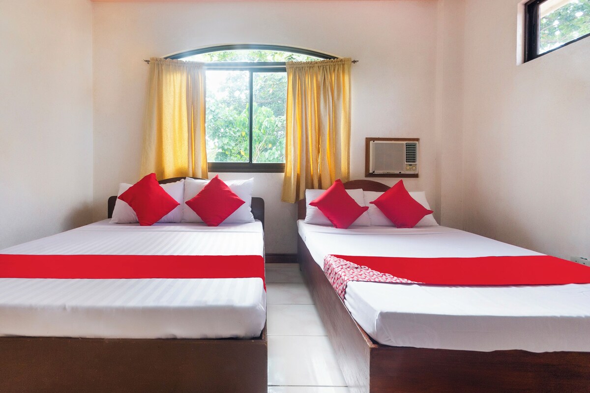 Habitación de hotel con dos camas, cada una con almohadas rojas y ropa de cama blanca, una ventana con cortinas amarillas y un equipo de aire acondicionado en la pared.