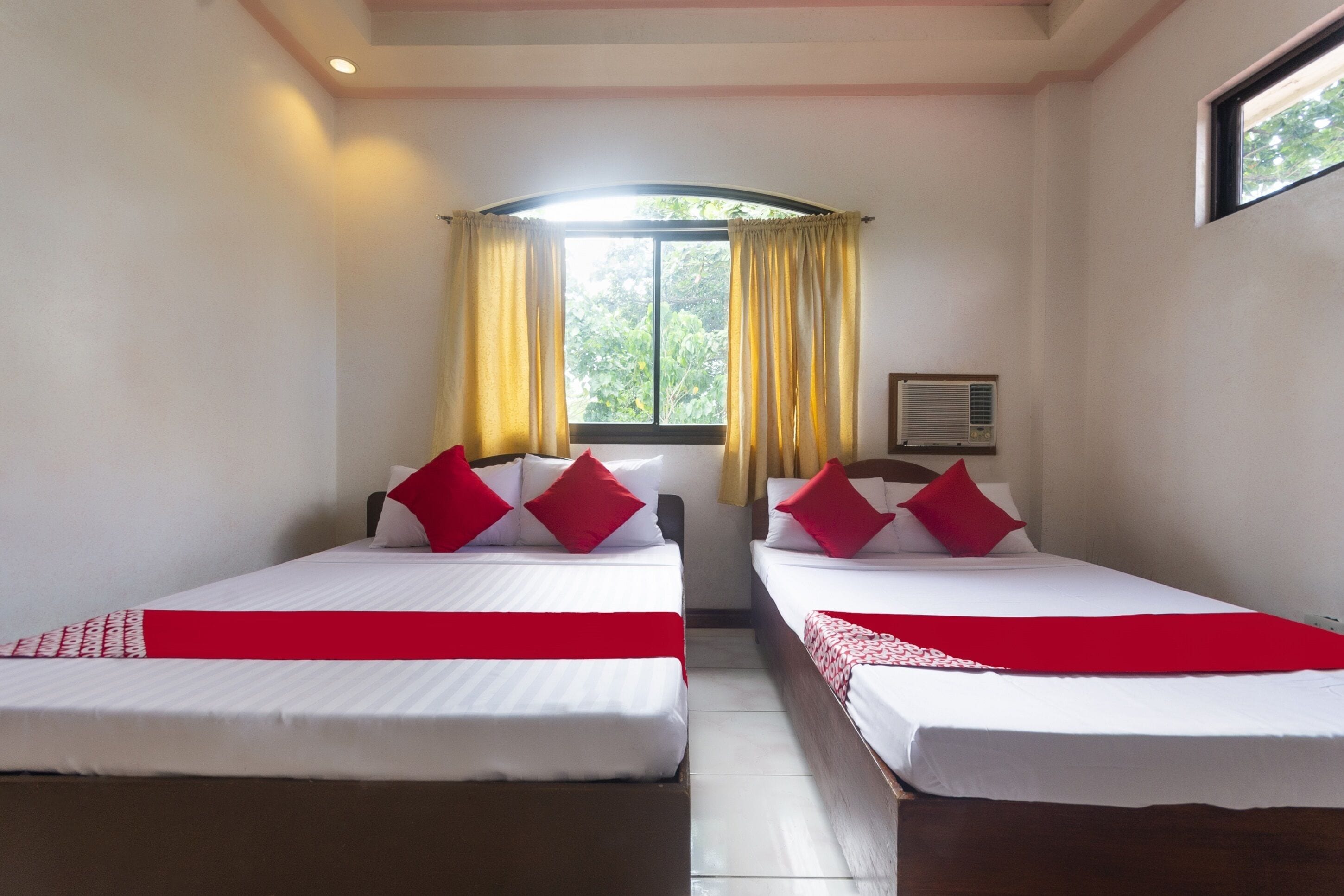 A hotel room with two beds, each with white linens and red accents, a window with yellow curtains, and a ceiling fan.