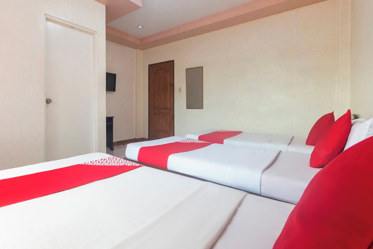 Habitación de hotel con tres camas dispuestas en fila, una puerta de madera, una mesita y un televisor montado en la pared.