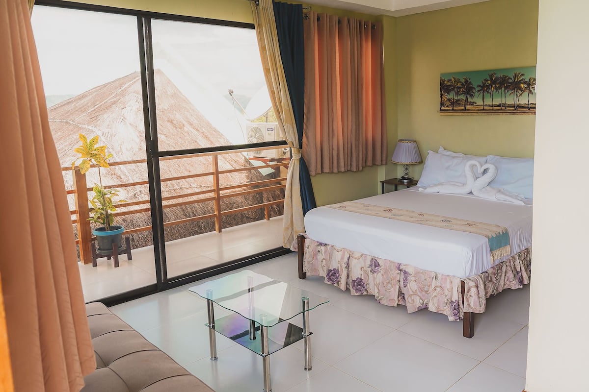 Habitación de hotel con cama, sofá, una mesita, ventilador de techo y un ventanal con cortinas.