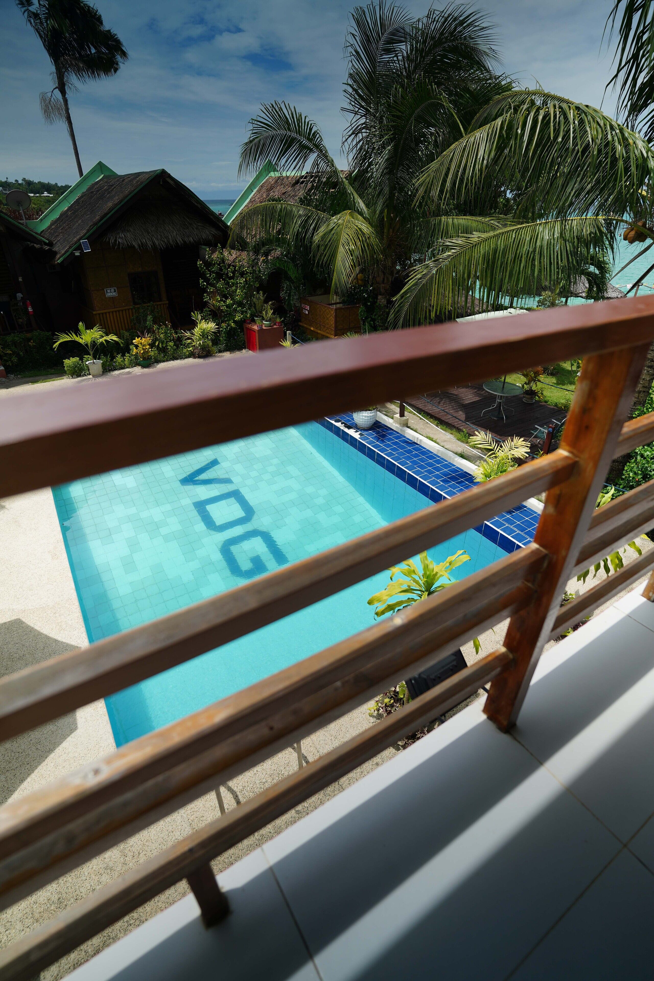 A pool with "VDG" written on it, surrounded by palm trees and a wooden deck.