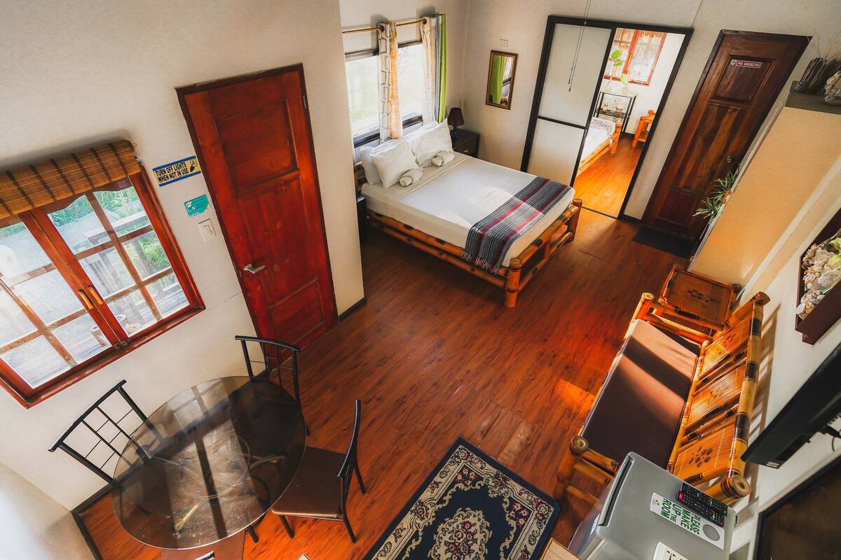 Una habitación con una cama, una mesa y una silla. Hay una ventana con cortinas, una puerta y un espejo en la pared.