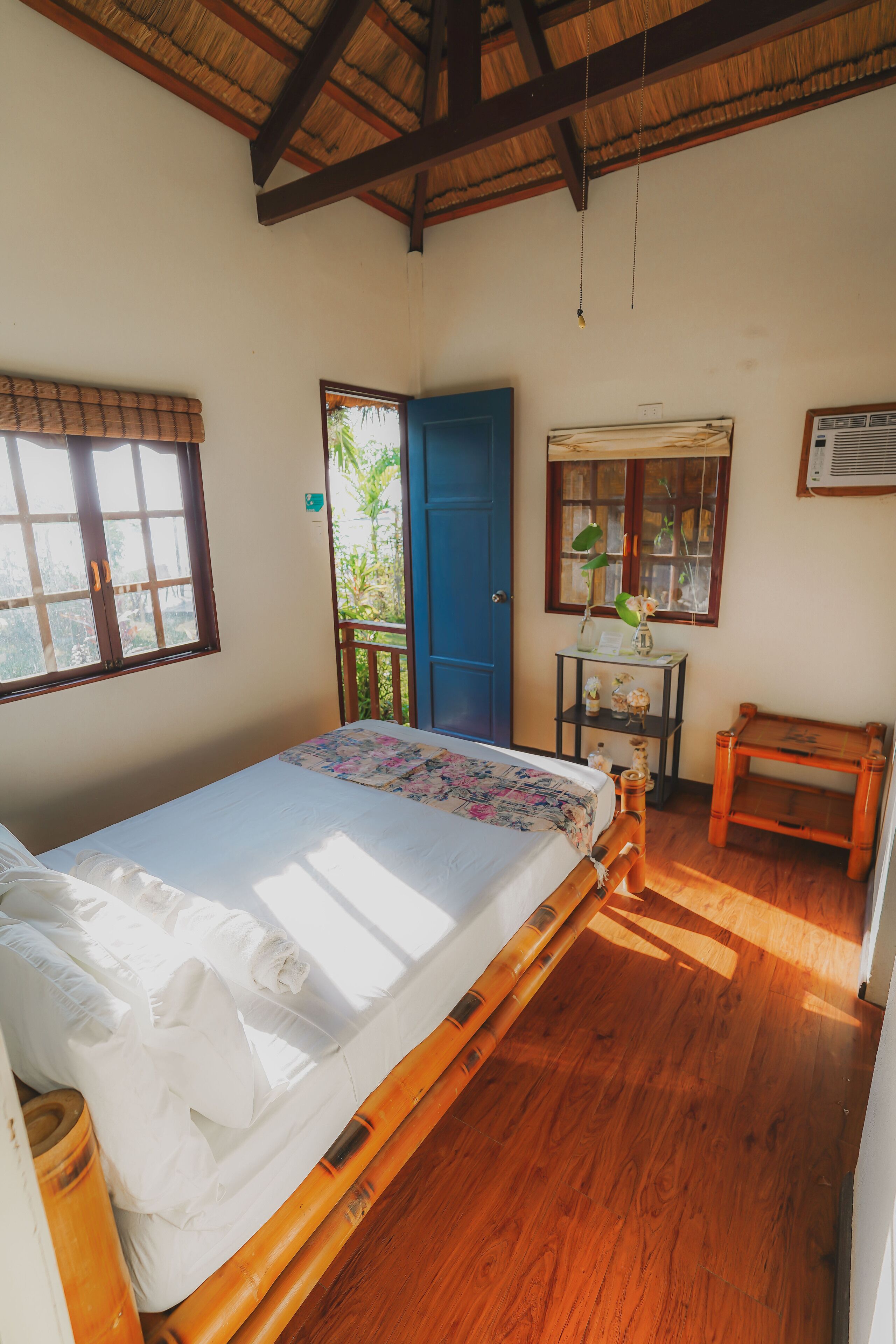 A room with a bed, a wooden desk, a small table, and a window with a view of greenery.