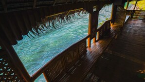 Una terraza de madera con vista a aguas azules cristalinas, barandillas de madera y una estructura de techo de paja.
