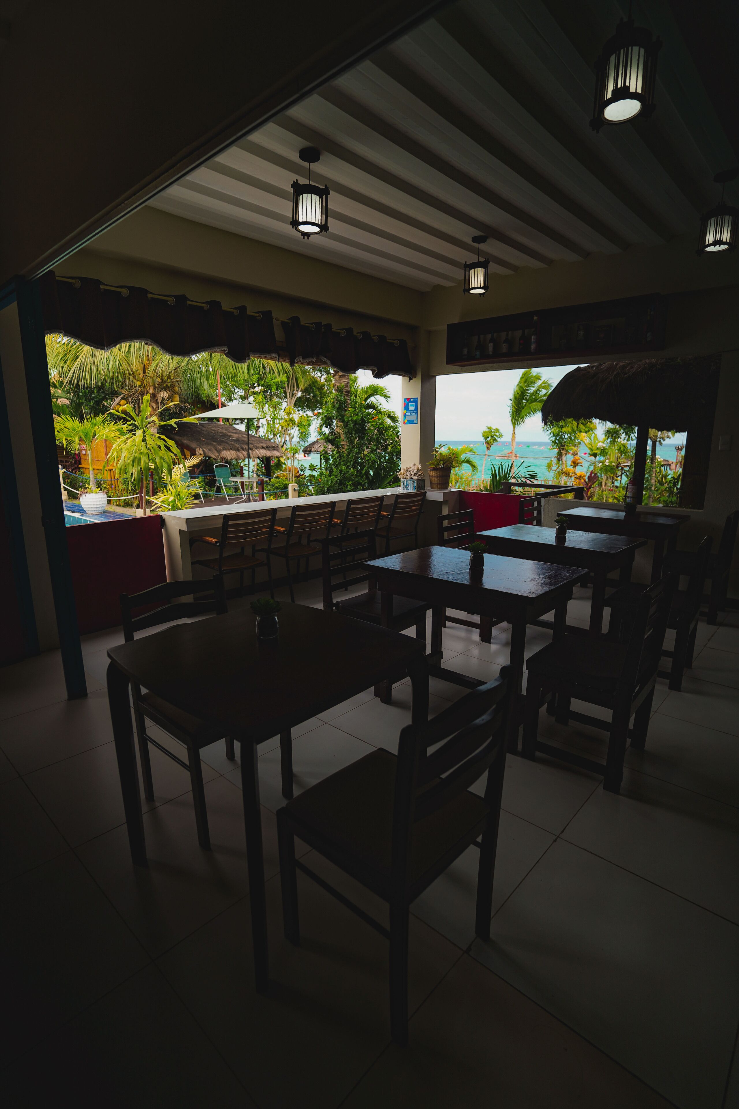 Un área de comedor al aire libre con mesas y sillas de madera, techo de paja y vistas al océano y vegetación tropical.