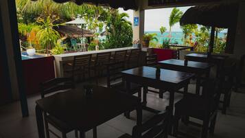 Un área de comedor al aire libre con mesas y sillas de madera, techo de paja y vistas al océano y vegetación tropical.
