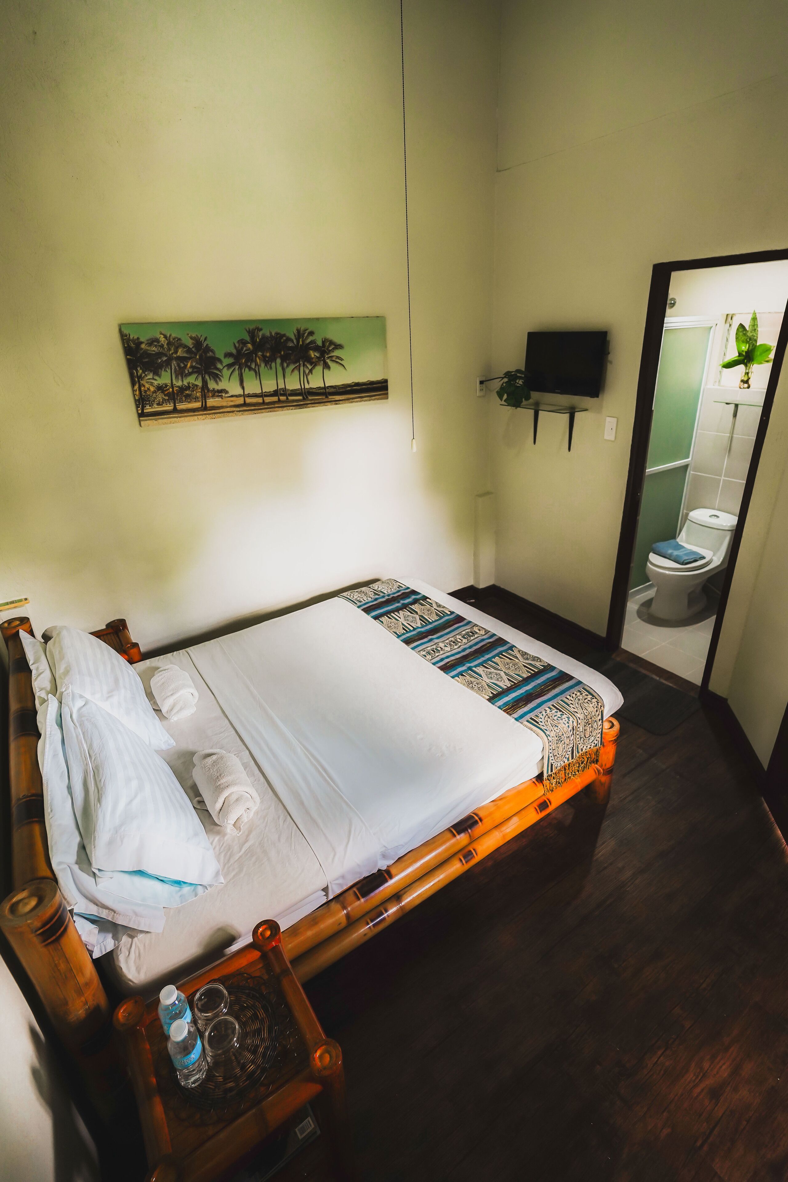 A neatly made bed with a patterned bedspread, a bedside table with a water bottle and glasses, a wall-mounted TV, and a painting of palm trees.