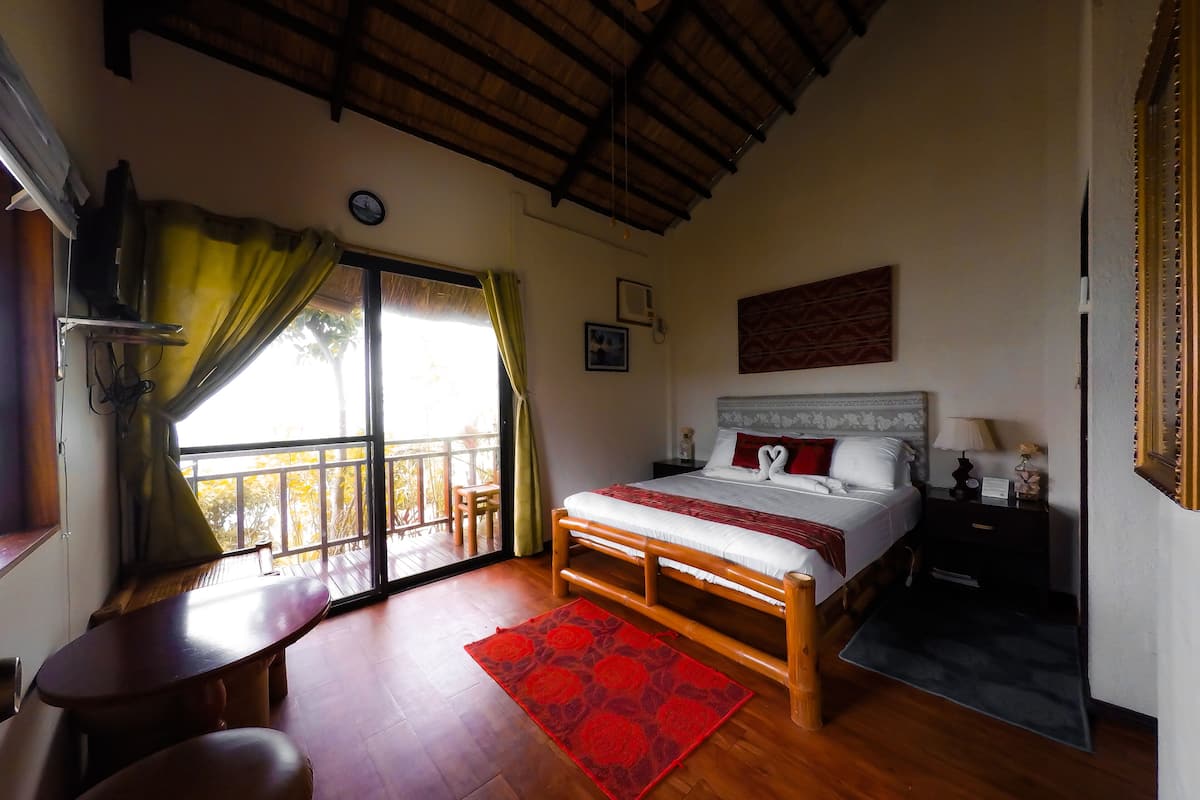 Un dormitorio con cama de madera, alfombra roja, mesita de noche, ventana con cortinas y balcón.