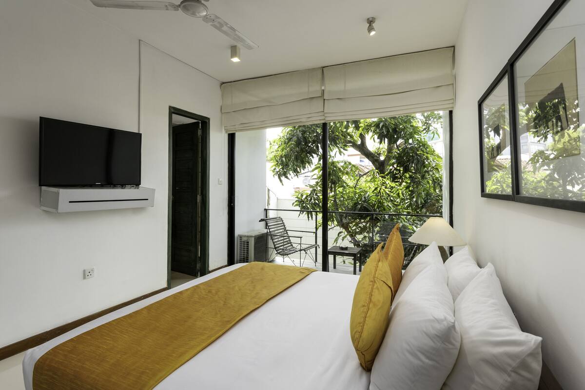 A hotel room with a bed, a mounted TV, a balcony with a chair, and a table.