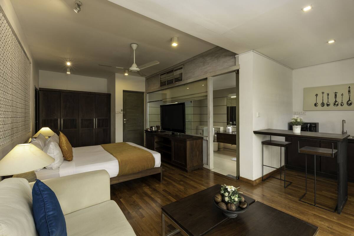 A modern hotel room with a bed, a sofa, a coffee table, a TV, a kitchenette, and a ceiling fan.