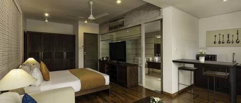 A modern hotel room with a bed, a sofa, a coffee table, a TV, a kitchenette, and a ceiling fan.