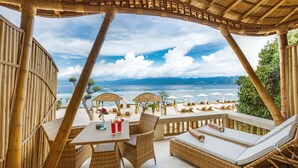 Un espacio de relax en la playa con mobiliario de mimbre, sombrillas y vistas al mar y a las montañas.