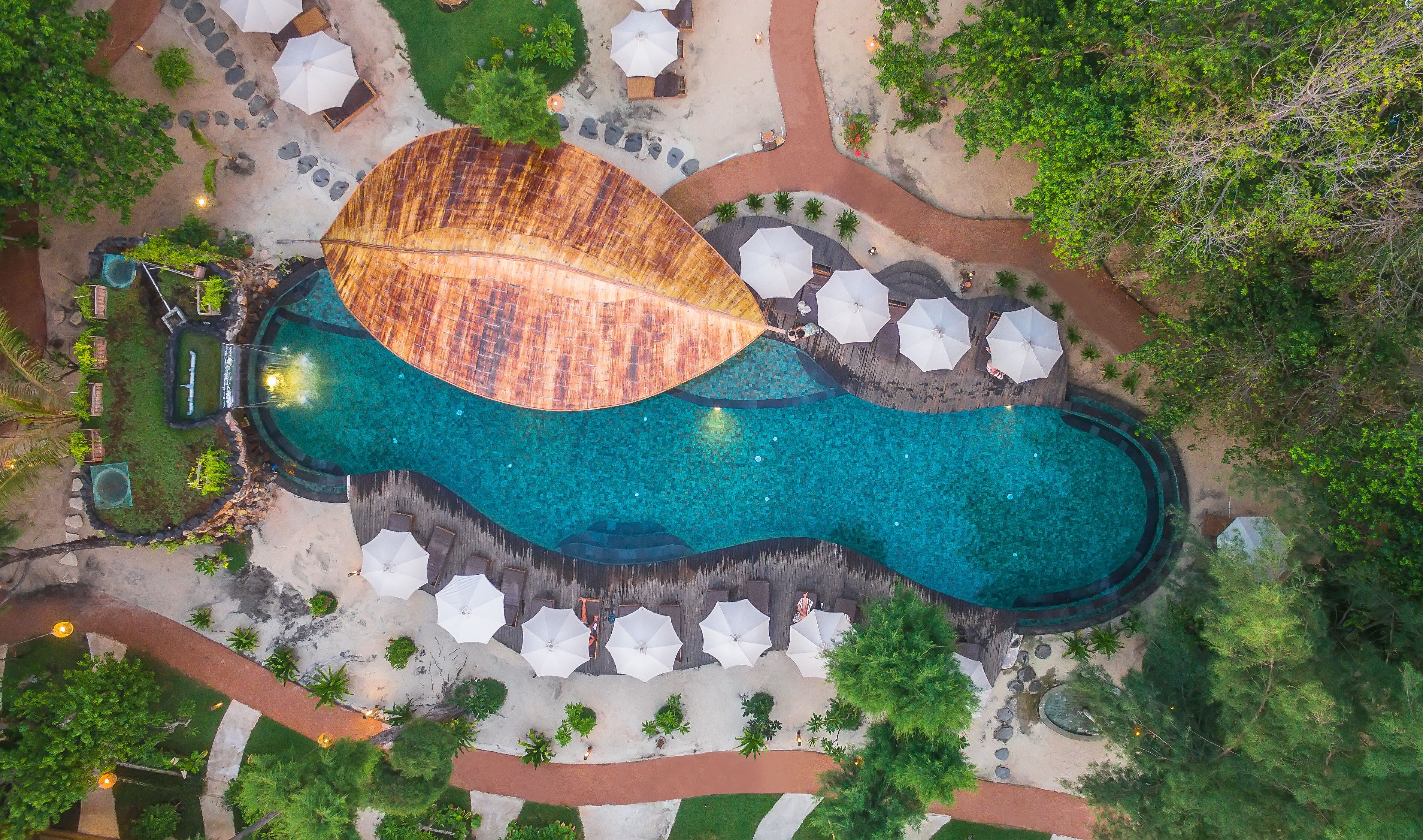 An aerial view of a resort with a large swimming pool, lounge areas, and a tropical garden.