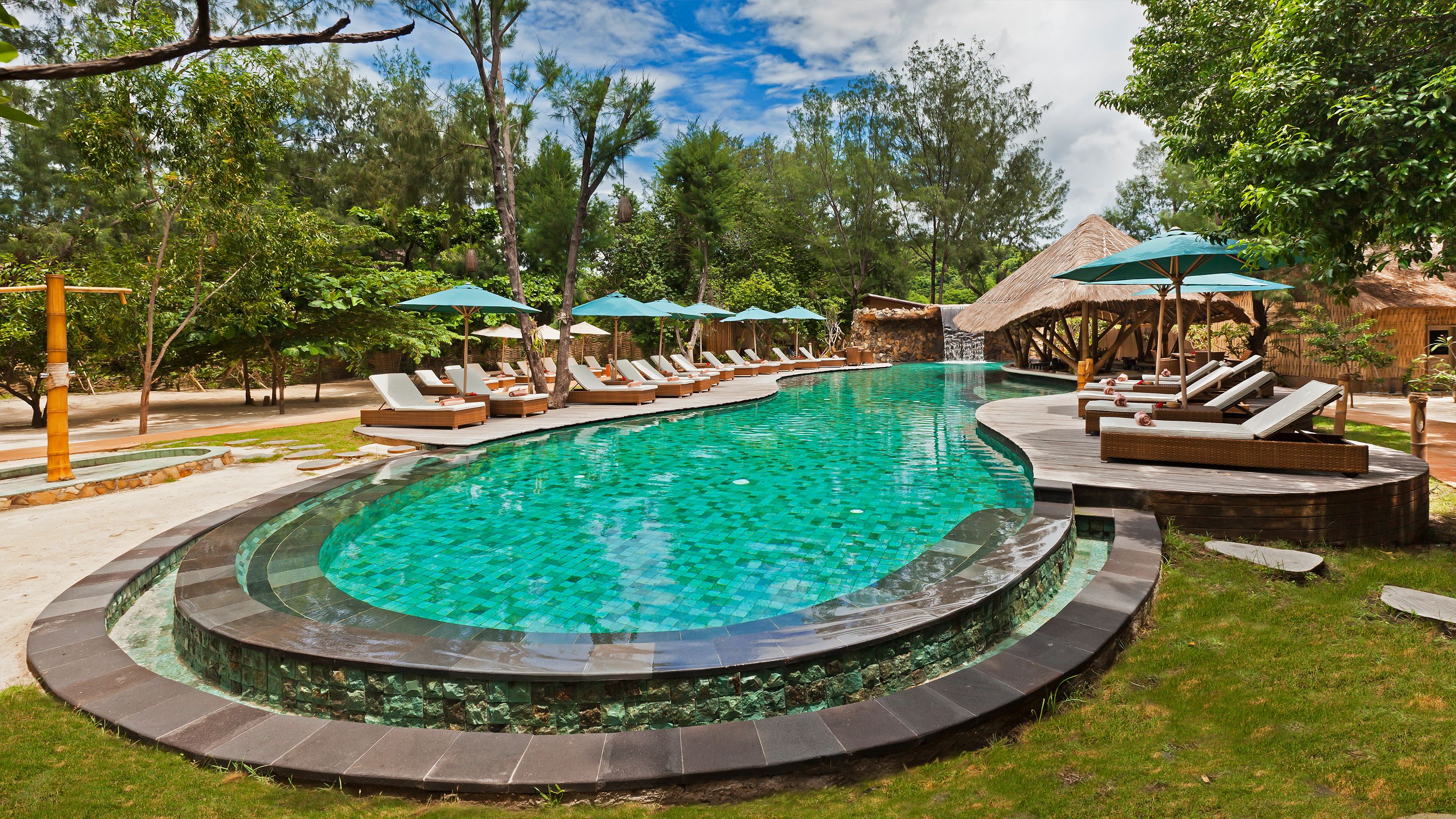 A large, curved swimming pool with lounge chairs and umbrellas, surrounded by trees and a thatched-roof structure.