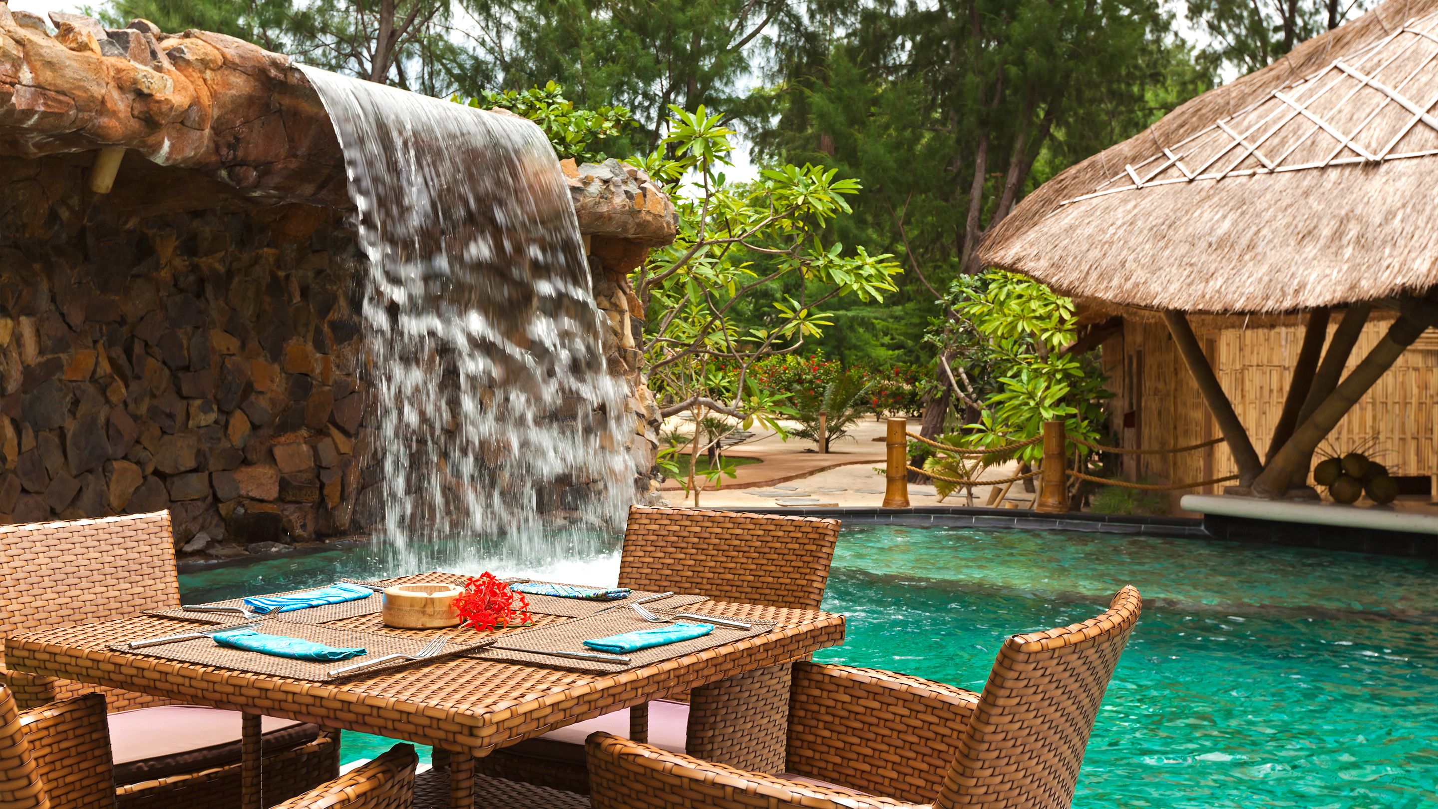 A pool area with a waterfall, wicker furniture, and a thatched-roof structure.