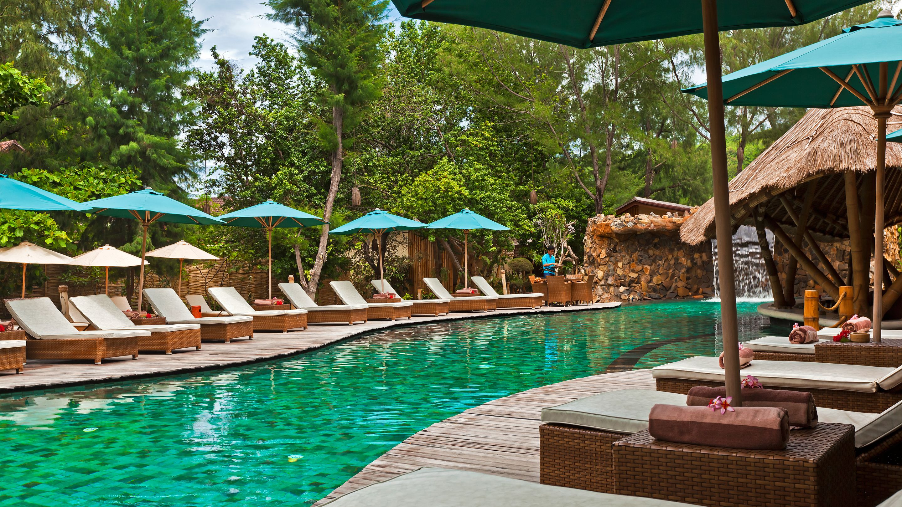 A poolside area with lounge chairs and umbrellas, a wooden deck, and a stone wall with a thatched roof.