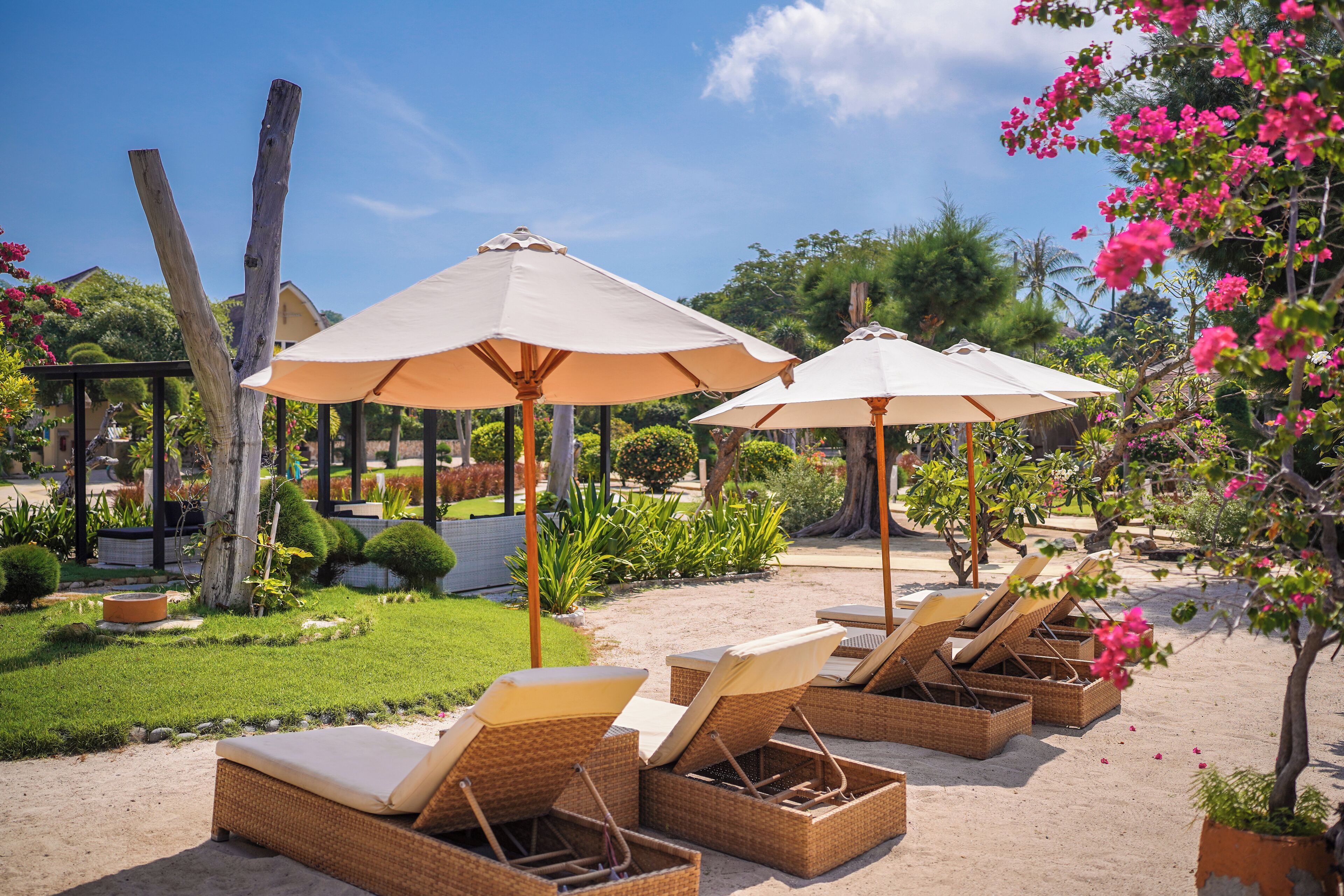 A beachside lounge area with wicker lounge chairs and beige umbrellas, surrounded by lush greenery and flowering plants.