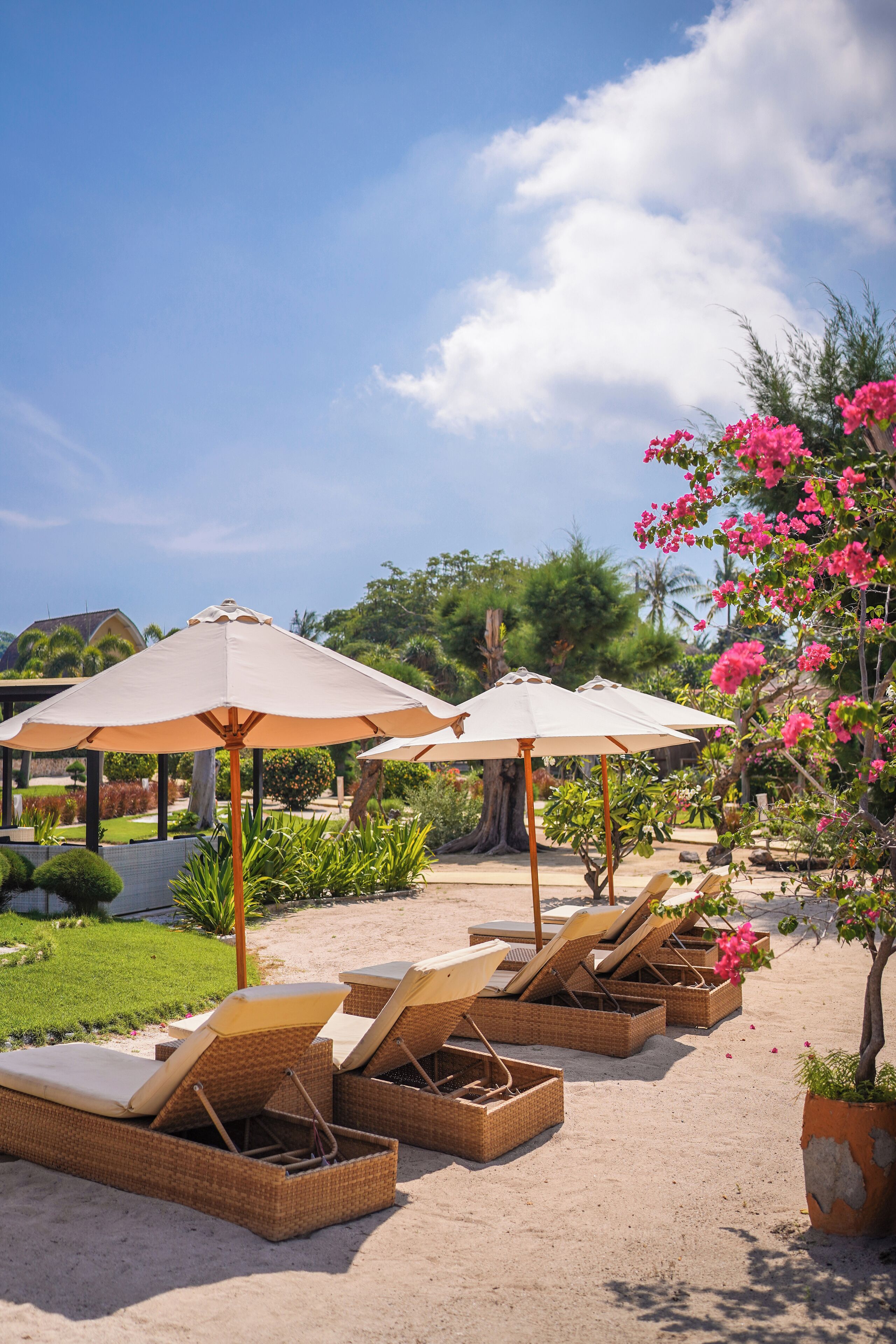 A beachside lounge area with wicker lounge chairs and beige umbrellas, surrounded by lush greenery and flowering plants.
