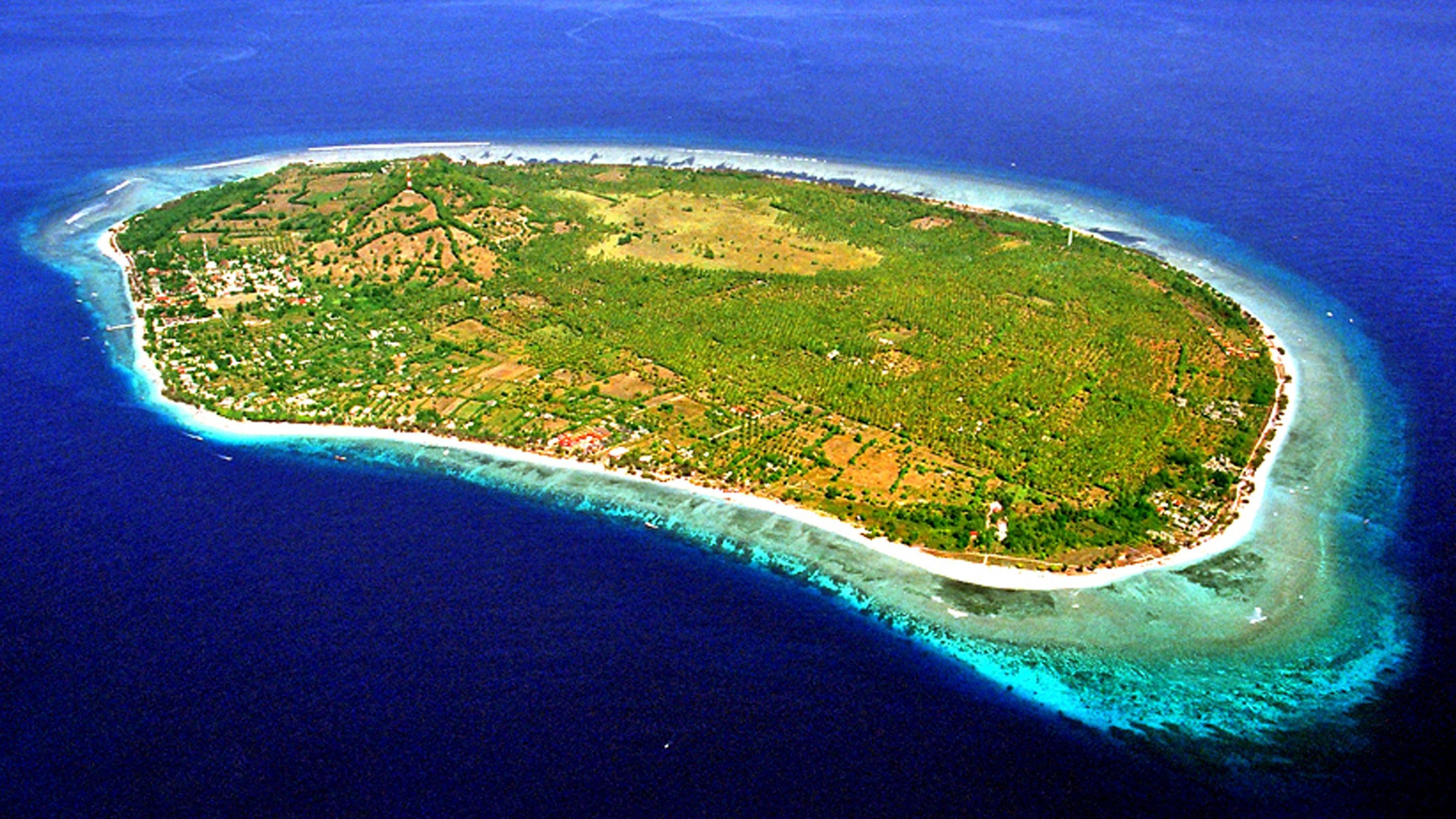 An aerial view of a small island with a narrow beach, surrounded by clear blue waters.