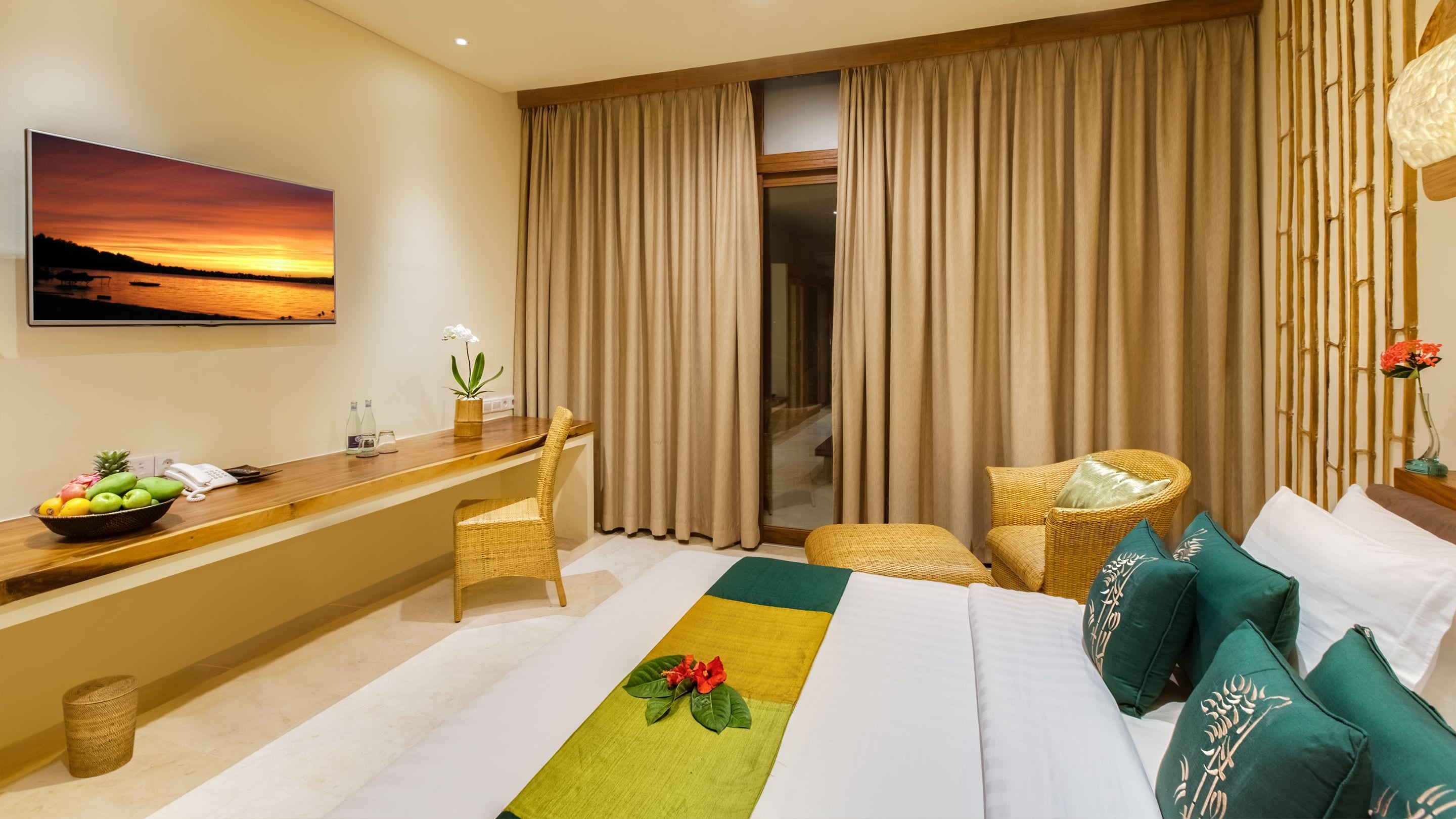 Une chambre d’hôtel avec un lit, un bureau sur lequel se trouve un panier de fruits, une chaise et une vue sur le coucher de soleil à travers les rideaux.