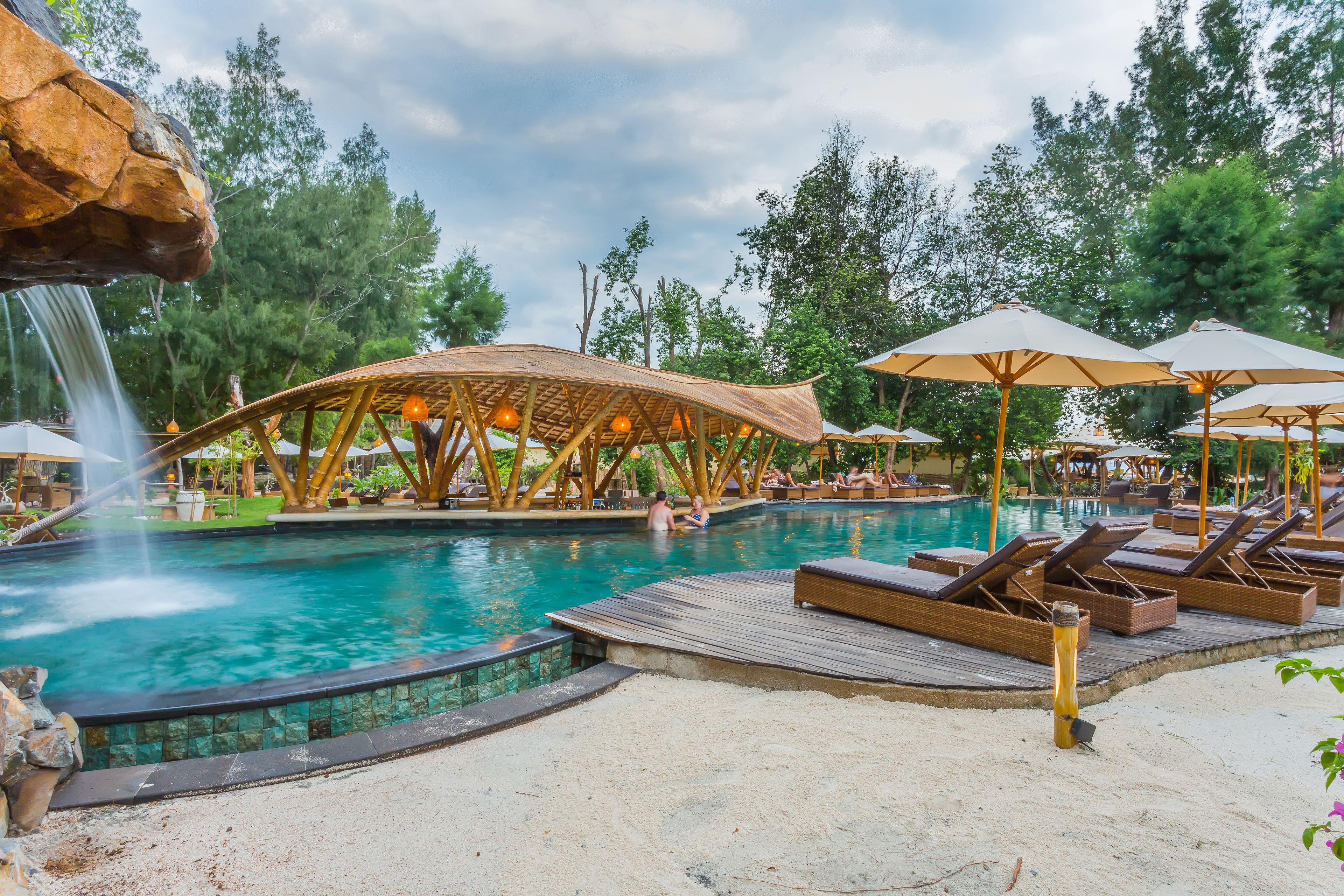 Zona de piscina con sillones de descanso y sombrillas, rodeada de árboles y una terraza de madera.