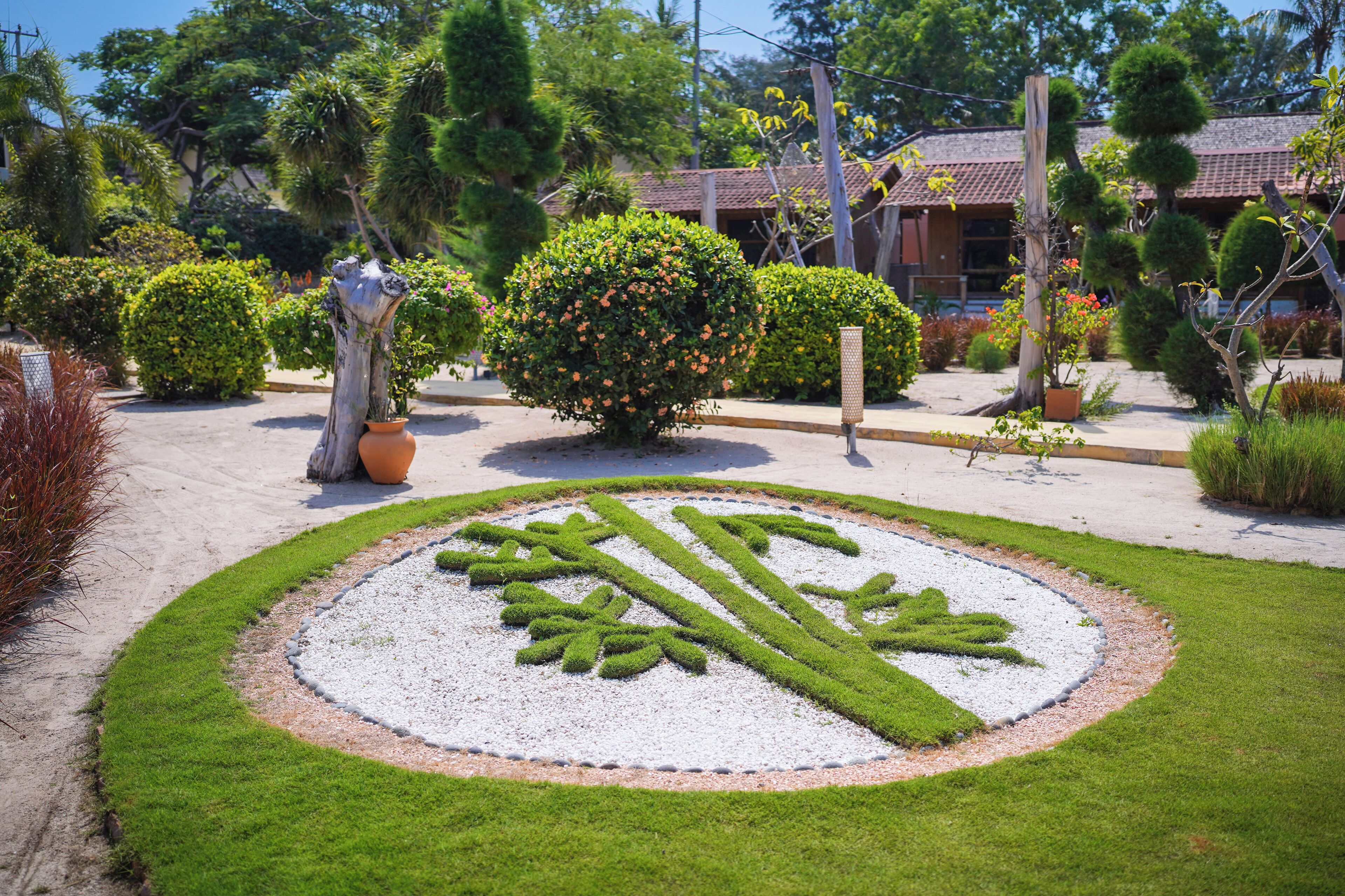 Um jardim ajardinado com design circular, com grama e cascalho, rodeado por arbustos e árvores diversos.