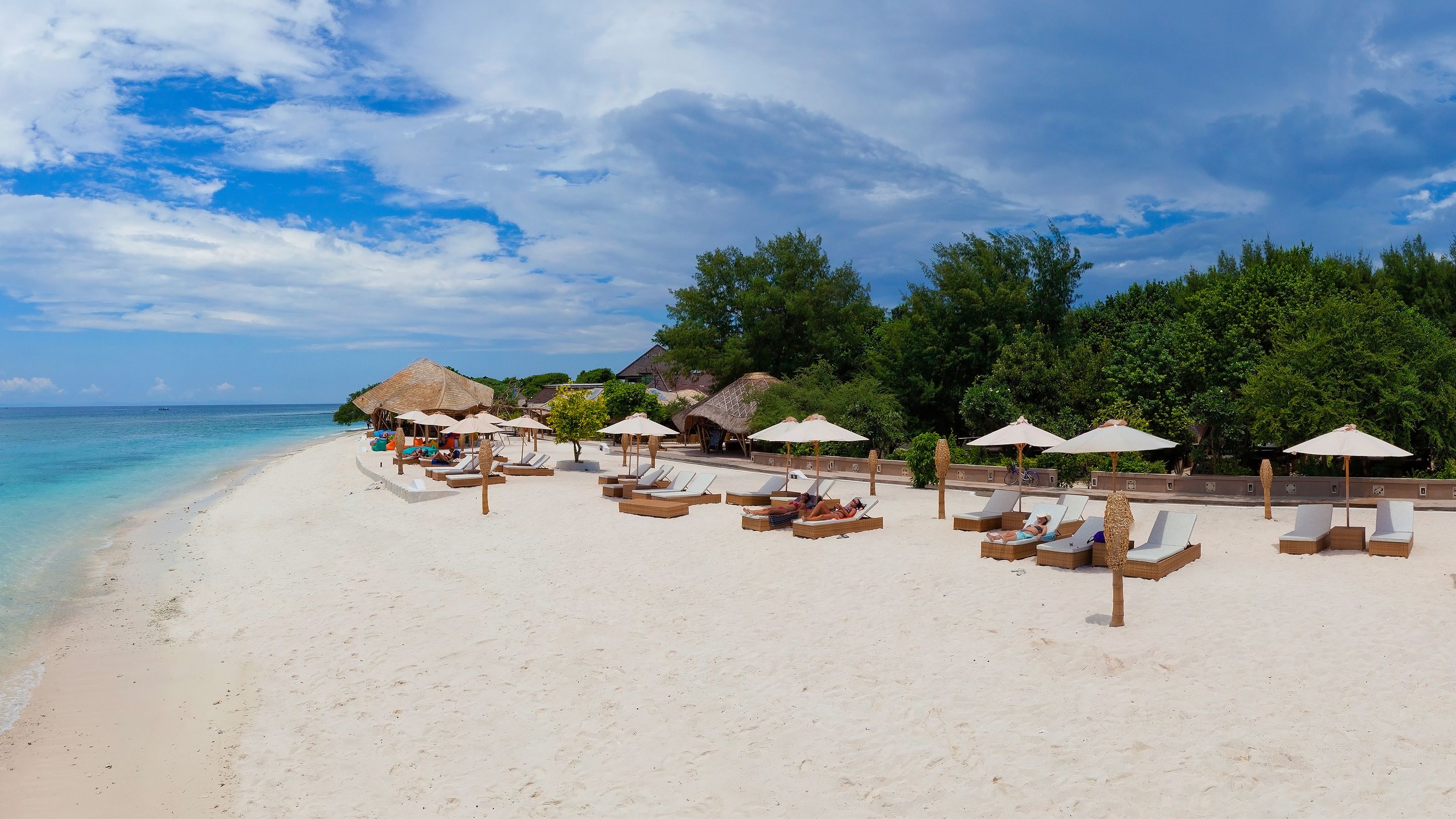 A beach with white sand, lounge chairs, umbrellas, and thatched-roof structures.