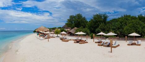 A beach with white sand, lounge chairs, umbrellas, and thatched-roof structures.