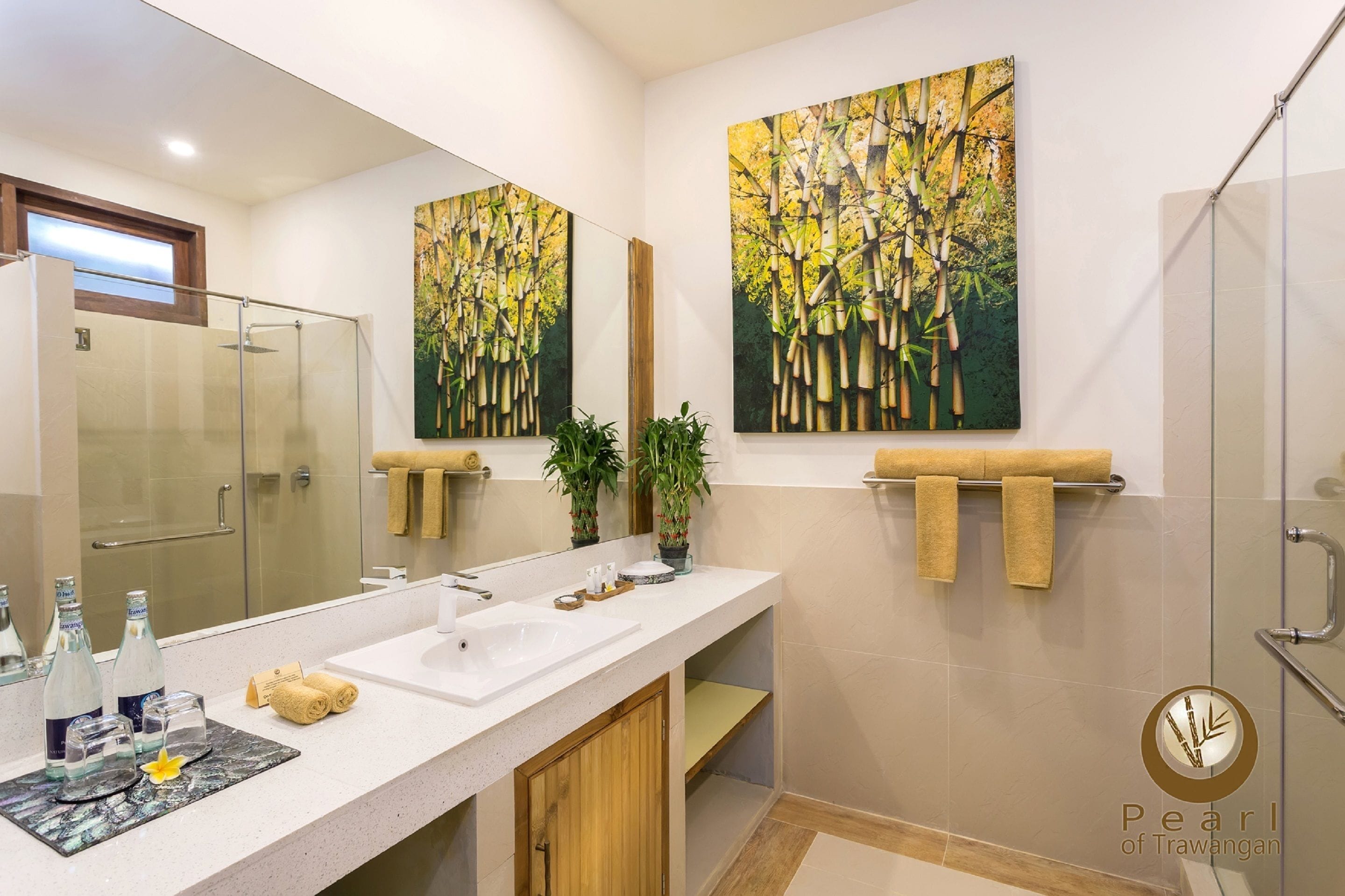 A modern bathroom with a large mirror, two sinks, and a glass shower enclosure. There are two large bamboo paintings on the wall, a towel rack, and a small plant.