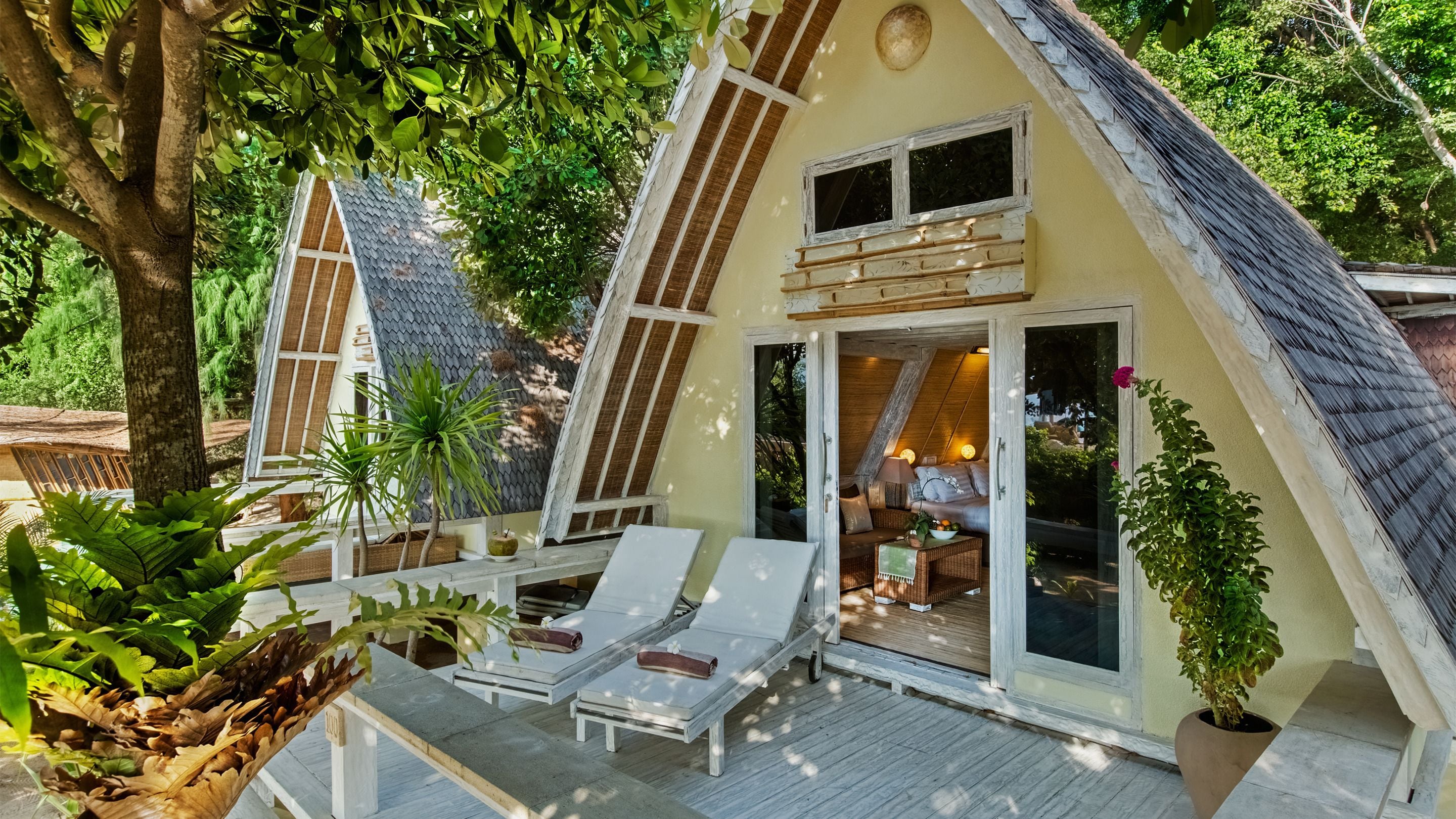 A cozy, triangular-roofed cabin with a deck, white lounge chairs, and a dining area with a table and chairs.