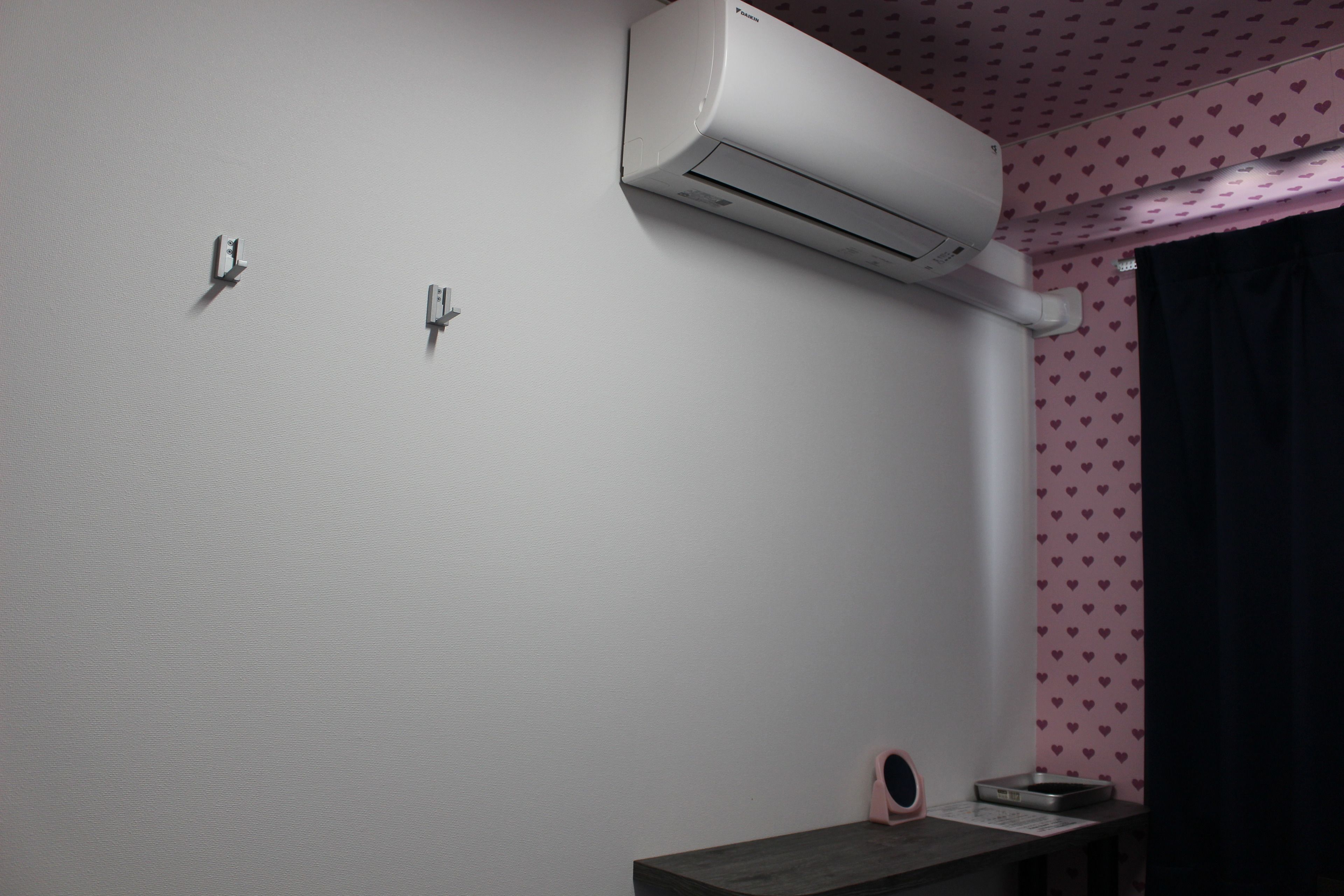 A room with a white wall, an air conditioning unit, a small table with a speaker, and a pink heart-patterned curtain.