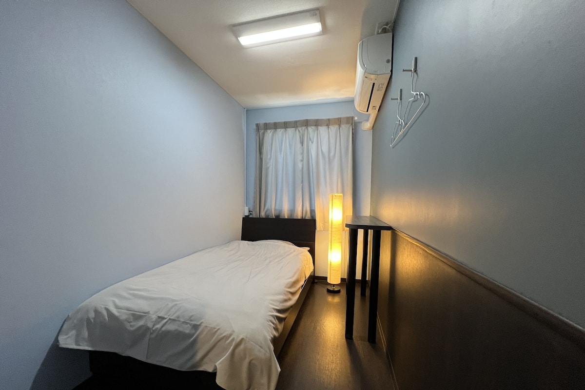 A hotel room with a bed, a bedside table with a lamp, a wall-mounted air conditioner, and a window with curtains.