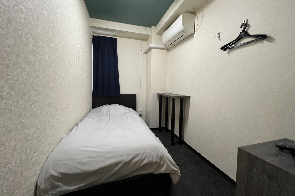 A small hotel room with a single bed, a bedside table, a hanging coat rack, and an air conditioning unit on the wall.