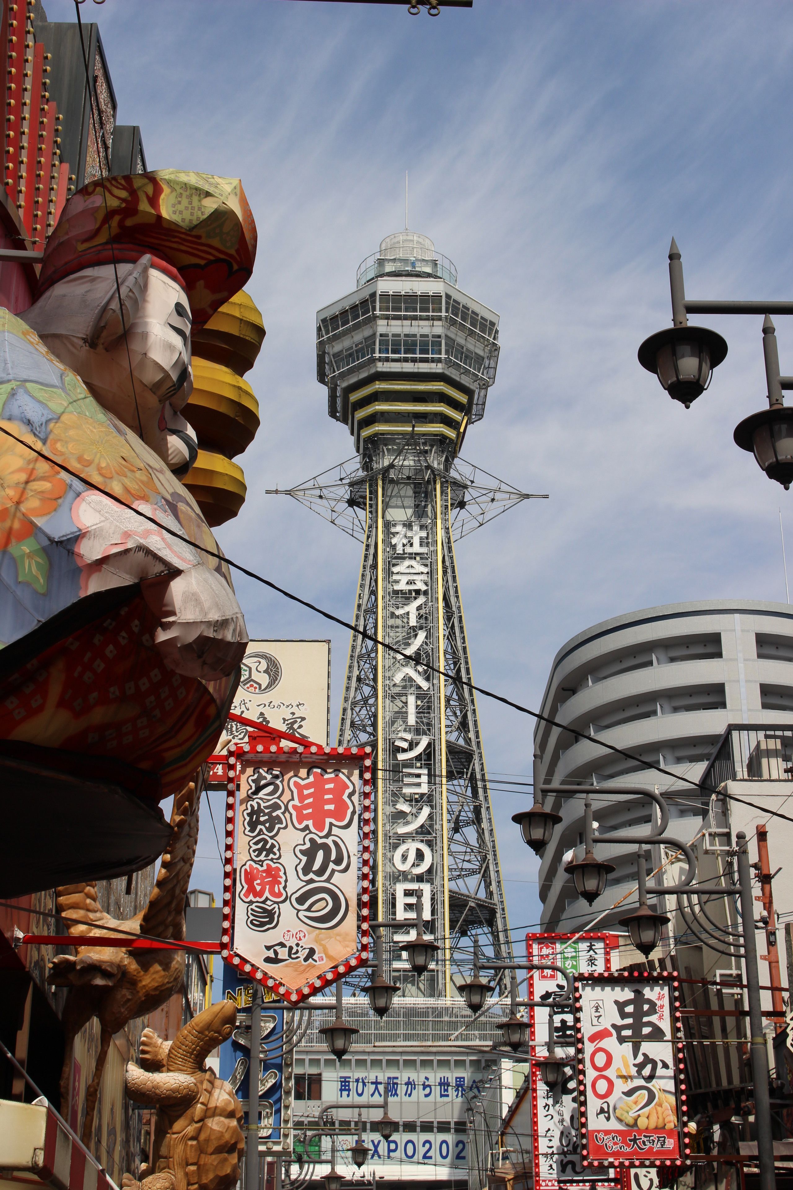 A large, colorful statue of a mythical creature, a tall tower with a unique design, and various signs and advertisements in a busy urban setting.
