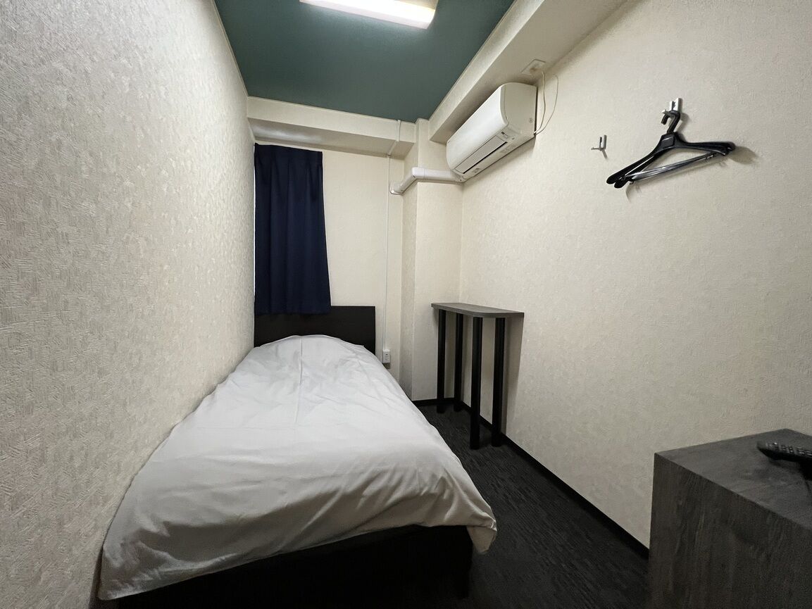 Ein kleines, schmales Hotelzimmer mit einem Bett, einem Nachttisch, einem Spiegel und einer Wand-Klimaanlage.