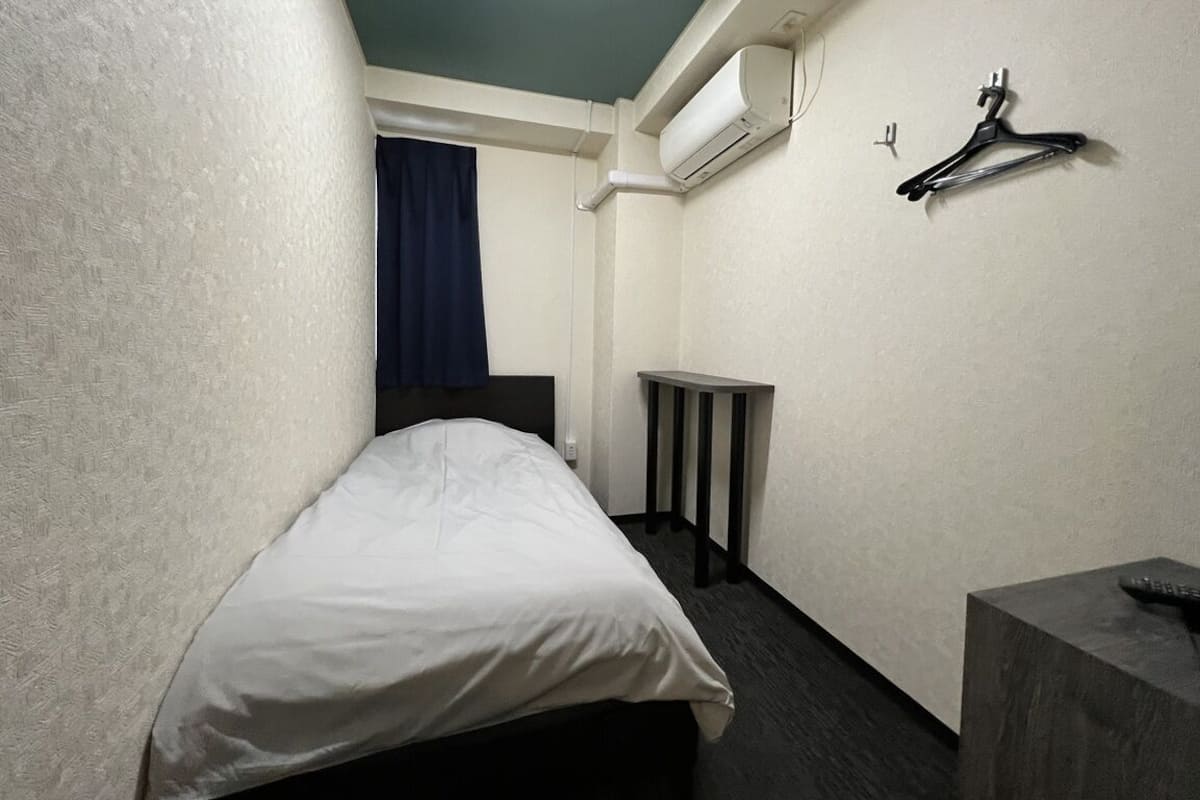 A small, narrow hotel room with a bed, a bedside table, a mirror, and a wall-mounted air conditioner.