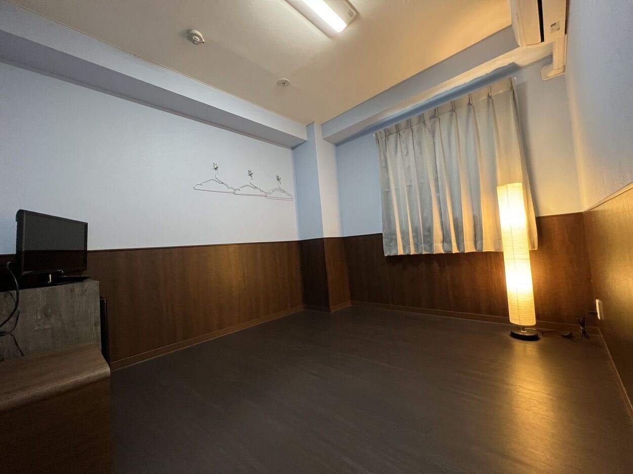 Ein Zimmer mit Holzboden, einem Flachbildfernseher auf einem Podest, einer Wandleuchte und einem Vorhangfenster.