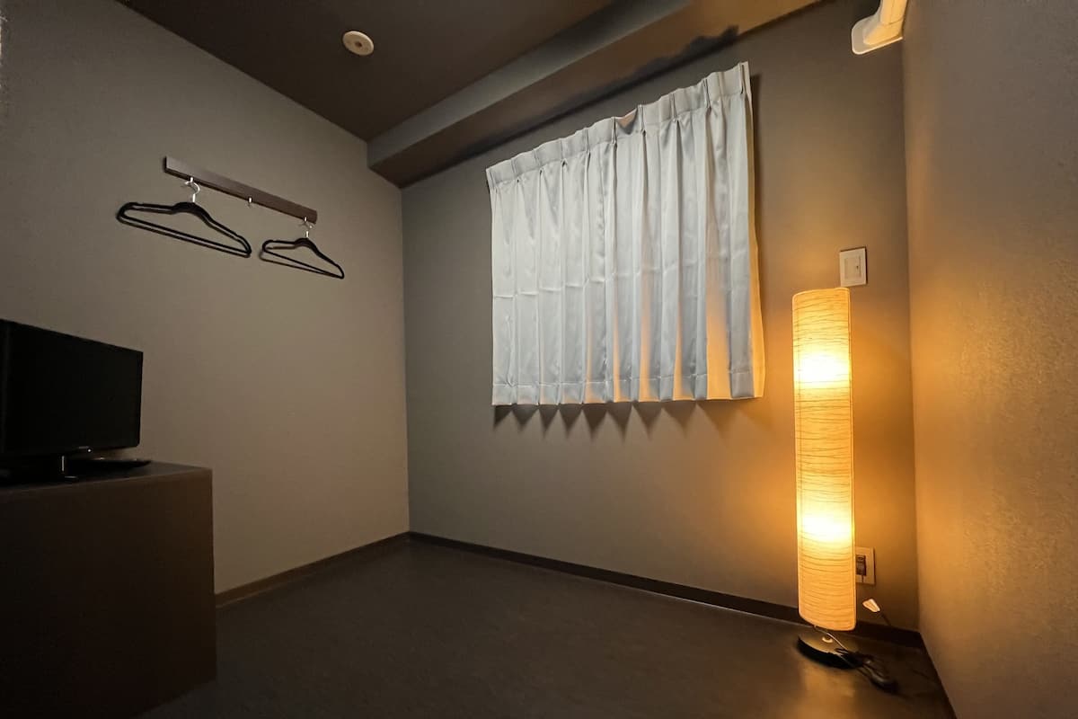 A hotel room with a flat-screen TV on a stand, a window with white curtains, a wall-mounted light fixture, and a coat rack with hangers.