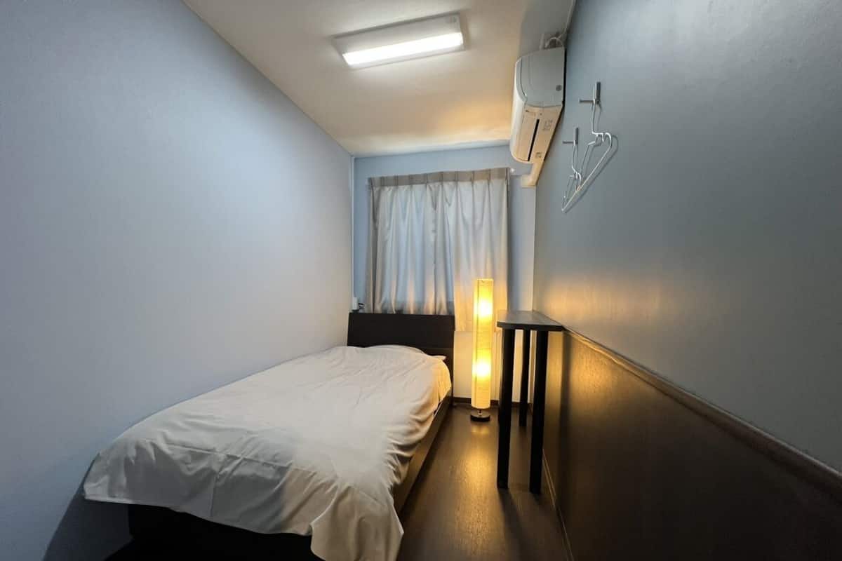 A hotel room with a bed, bedside table, lamp, air conditioning unit, and a window with curtains.