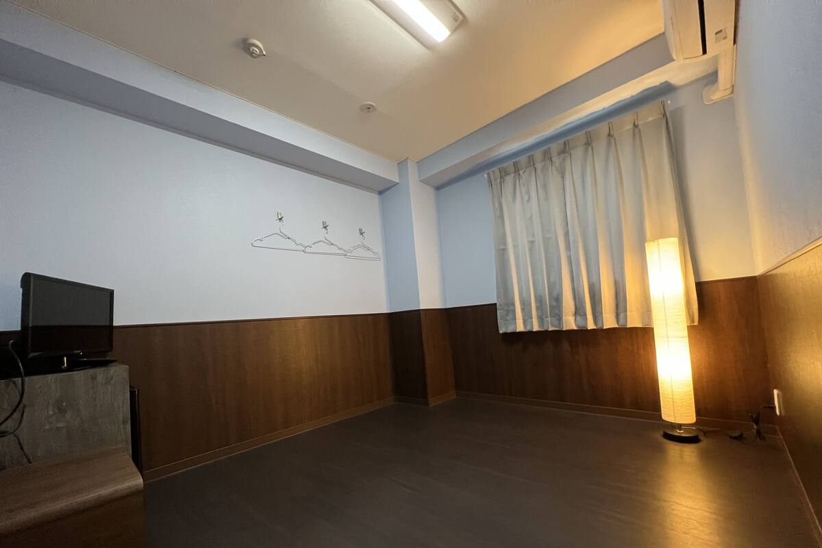 A room with a wooden floor, a flat-screen TV on a stand, a wall with a simple line drawing, and a large window with curtains.