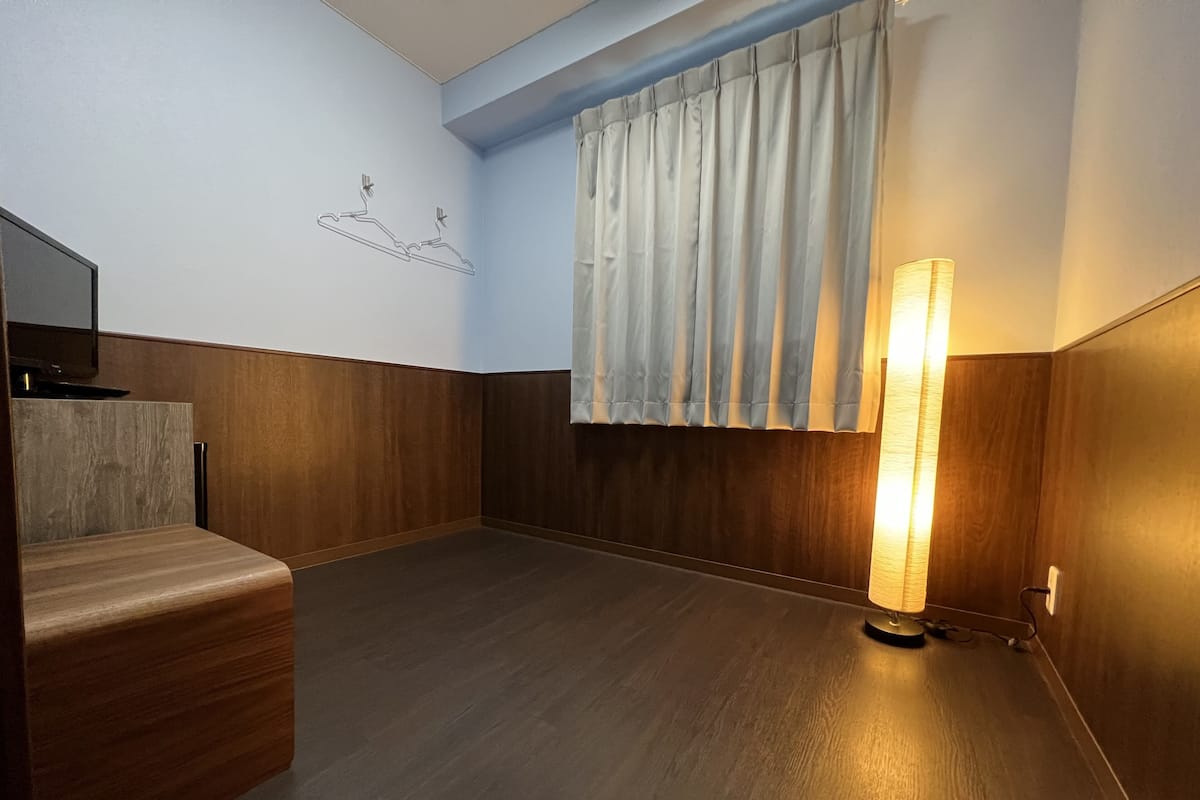 A hotel room with a wooden floor, a flat-screen TV mounted on the wall, a standing lamp, and a window with curtains.
