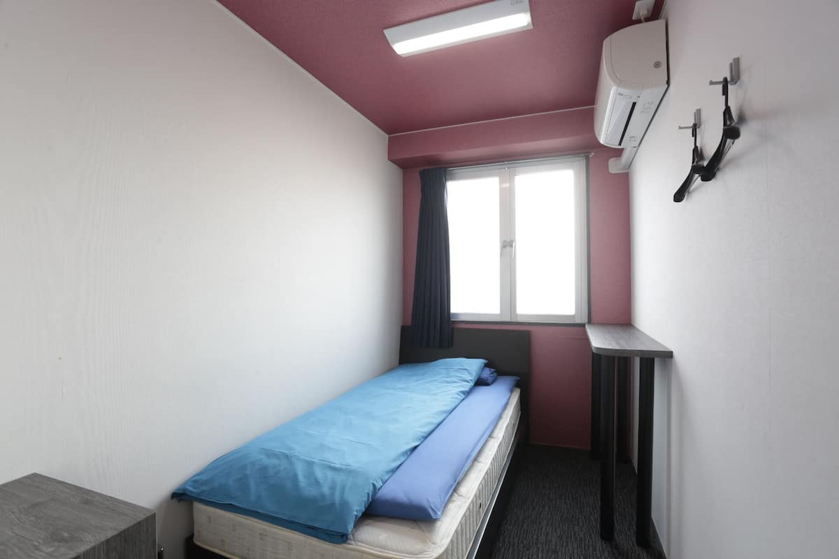 A small, single-bed room with a window, a desk, and an air conditioning unit.