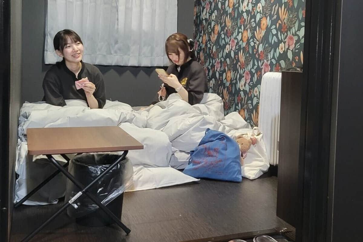 Two people sitting on a bed in a room with a floral wallpaper, a small table, and a trash bin.