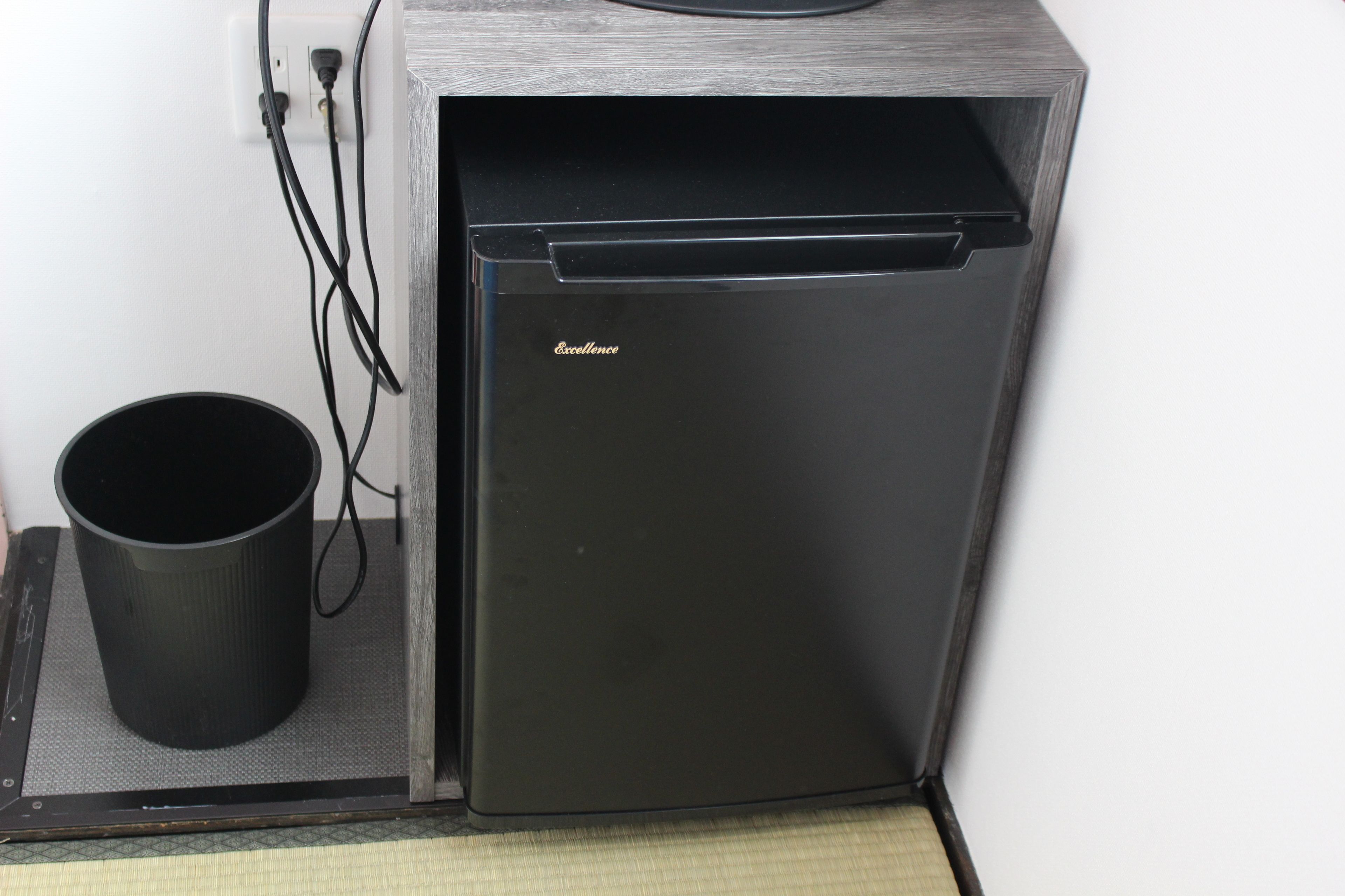 Un petit réfrigérateur noir, avec le nom de marque « Excellence » sur la porte, une tasse noire posée sur un tapis à côté, et un câble d’alimentation branché dans une prise murale.