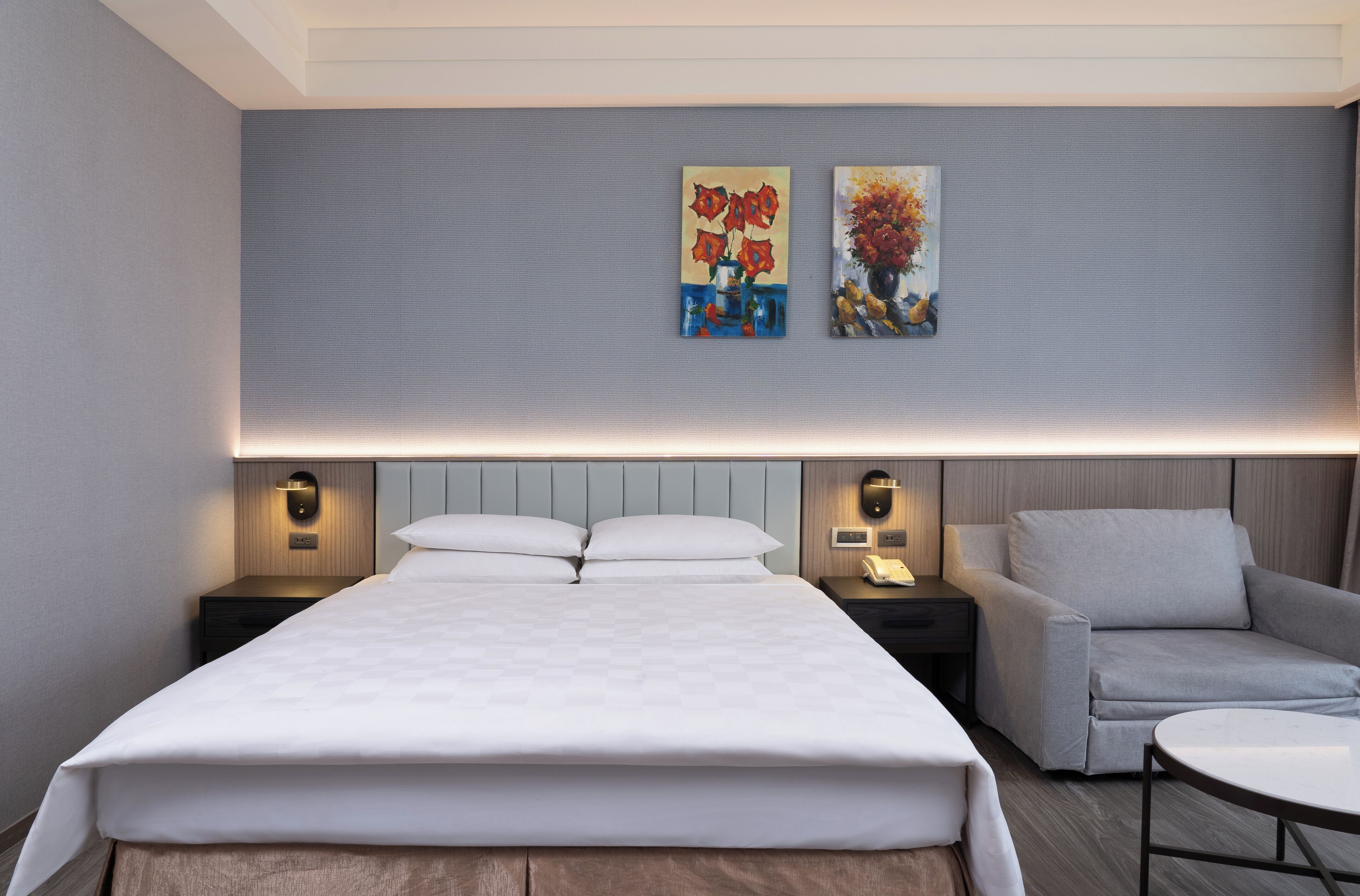 Una habitación de hotel moderna con una cama grande, dos mesitas de noche con lámparas, un sofá gris y dos cuadros en la pared.