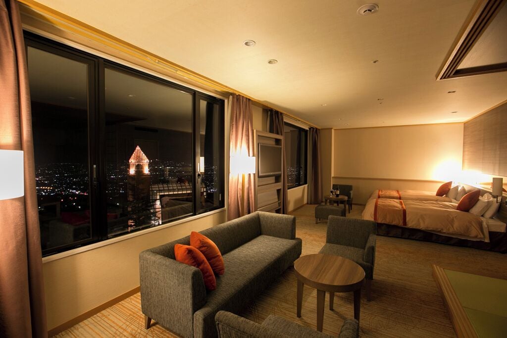 Een hotelkamer met een groot raam met uitzicht op de stad, een bank, een klein tafeltje en een bed met kussens.