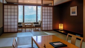 A traditional Japanese room with tatami flooring, a low table, chairs, a sliding door with a city view, and a framed picture on the wall. - Fruit Park Fujiya Hotel (Yamanashi)