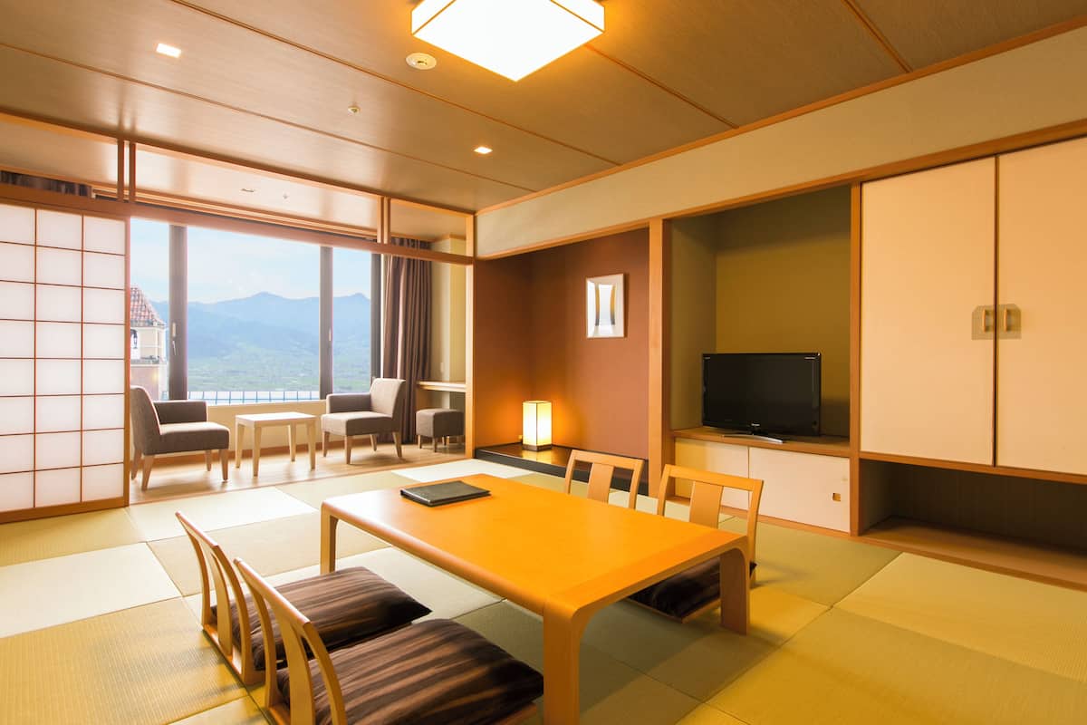 Ein traditioneller japanischer Raum mit Tatami-Böden, einem niedrigen Tisch mit Kissen, einem Fernseher und Bergblick durch große Fenster.