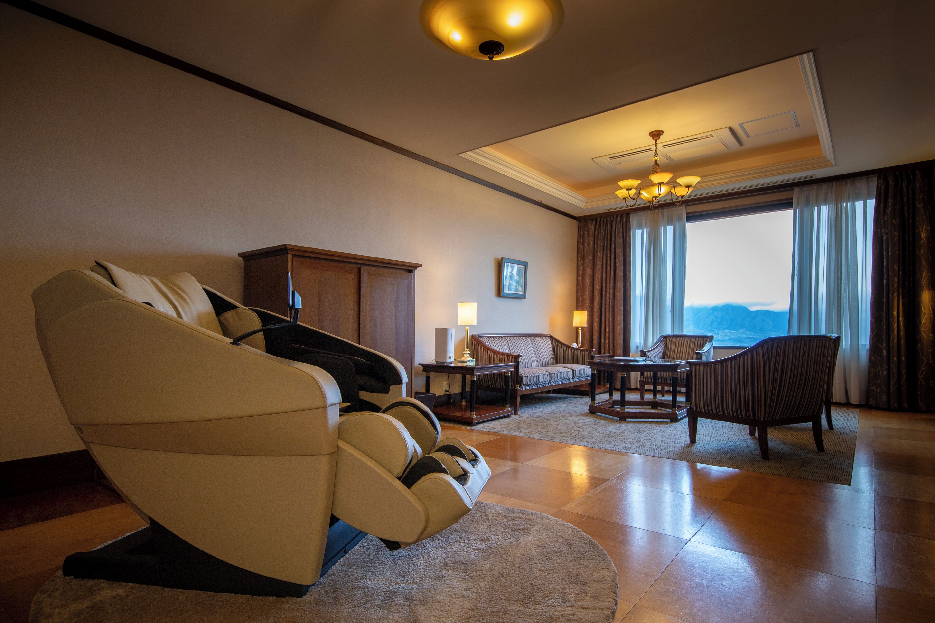 A spacious living room with a massage chair, a sofa, a dining table, and a view of the mountains through large windows.