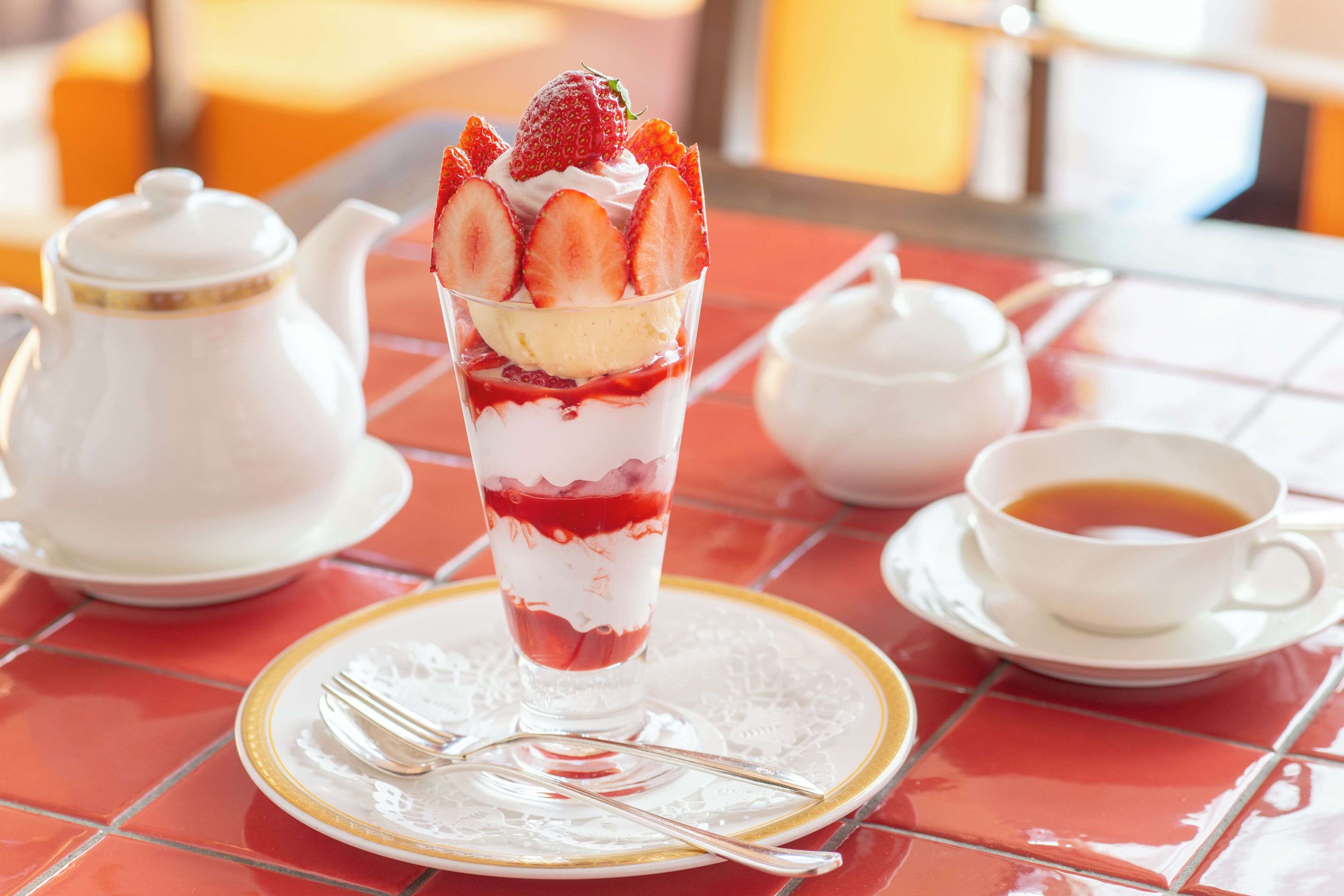 Een dessert met aardbeien en room in een glas, een theepot, een kop thee en een vork op een rode betegelde tafel.